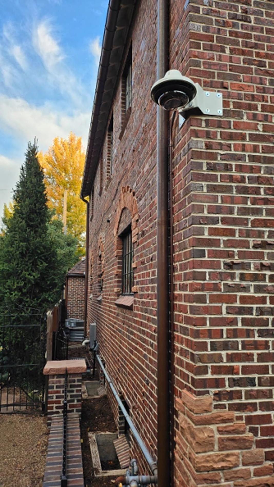 Brick building exterior with wall-mounted light, narrow side walkway, and trees in autumn colors.