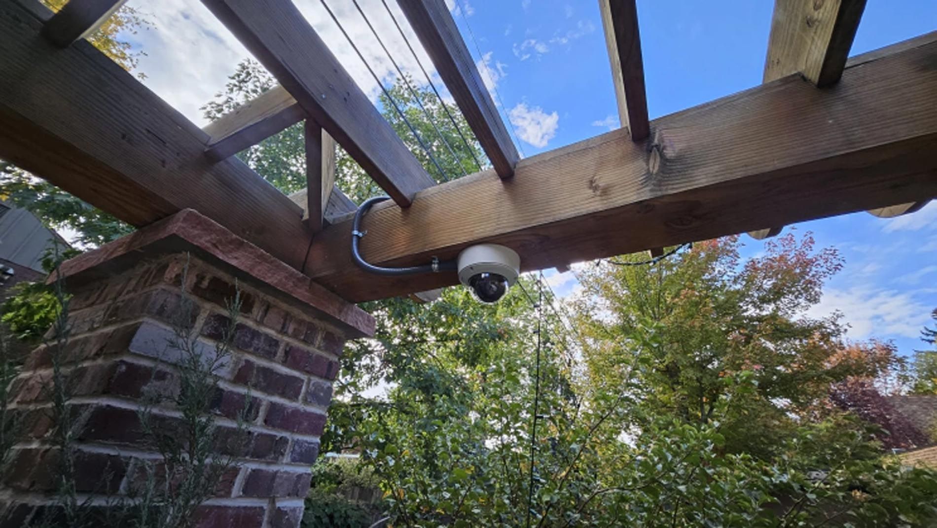 Security camera mounted under a wooden pergola beside a brick wall, overlooking a leafy yard