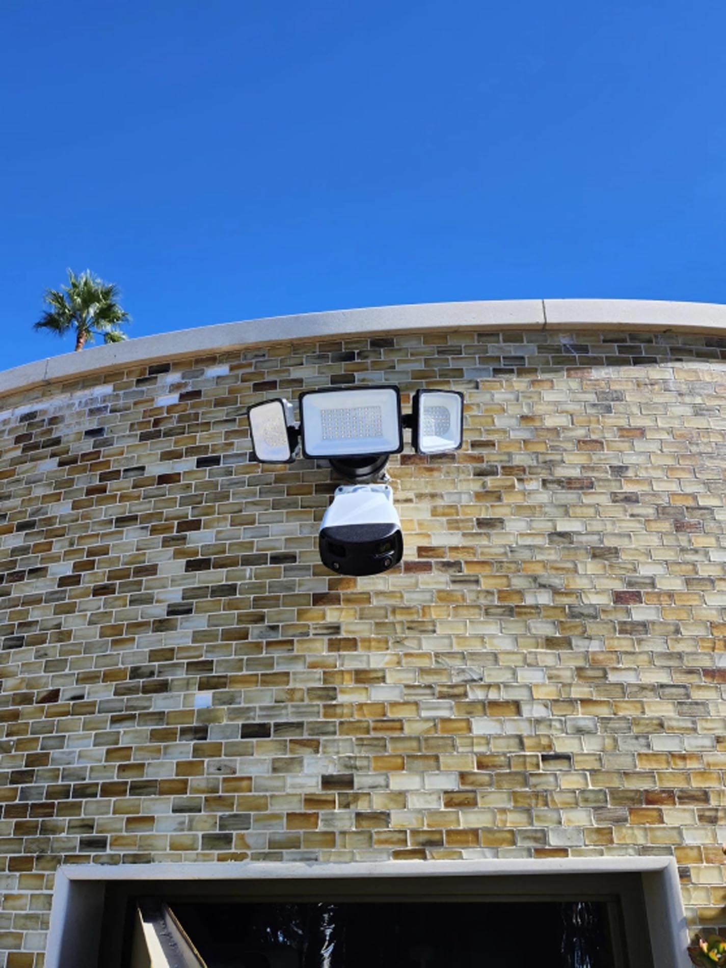 Outdoor security light mounted on a tan brick wall beneath a blue sky