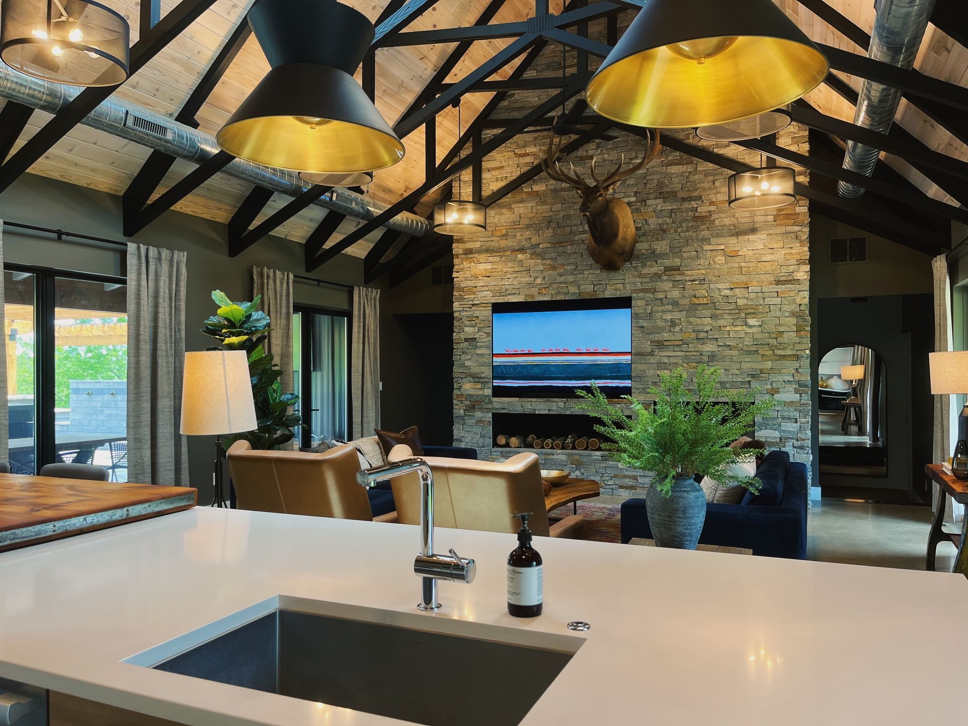 Modern living room with exposed beams, stone fireplace, large windows, and a kitchen island in the foreground
