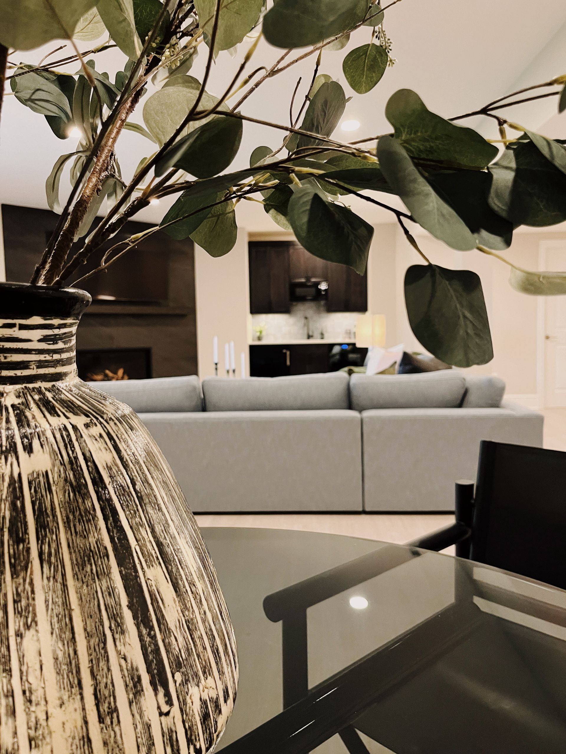 Vase with leafy branches on a table, with a gray sofa and kitchen in the background