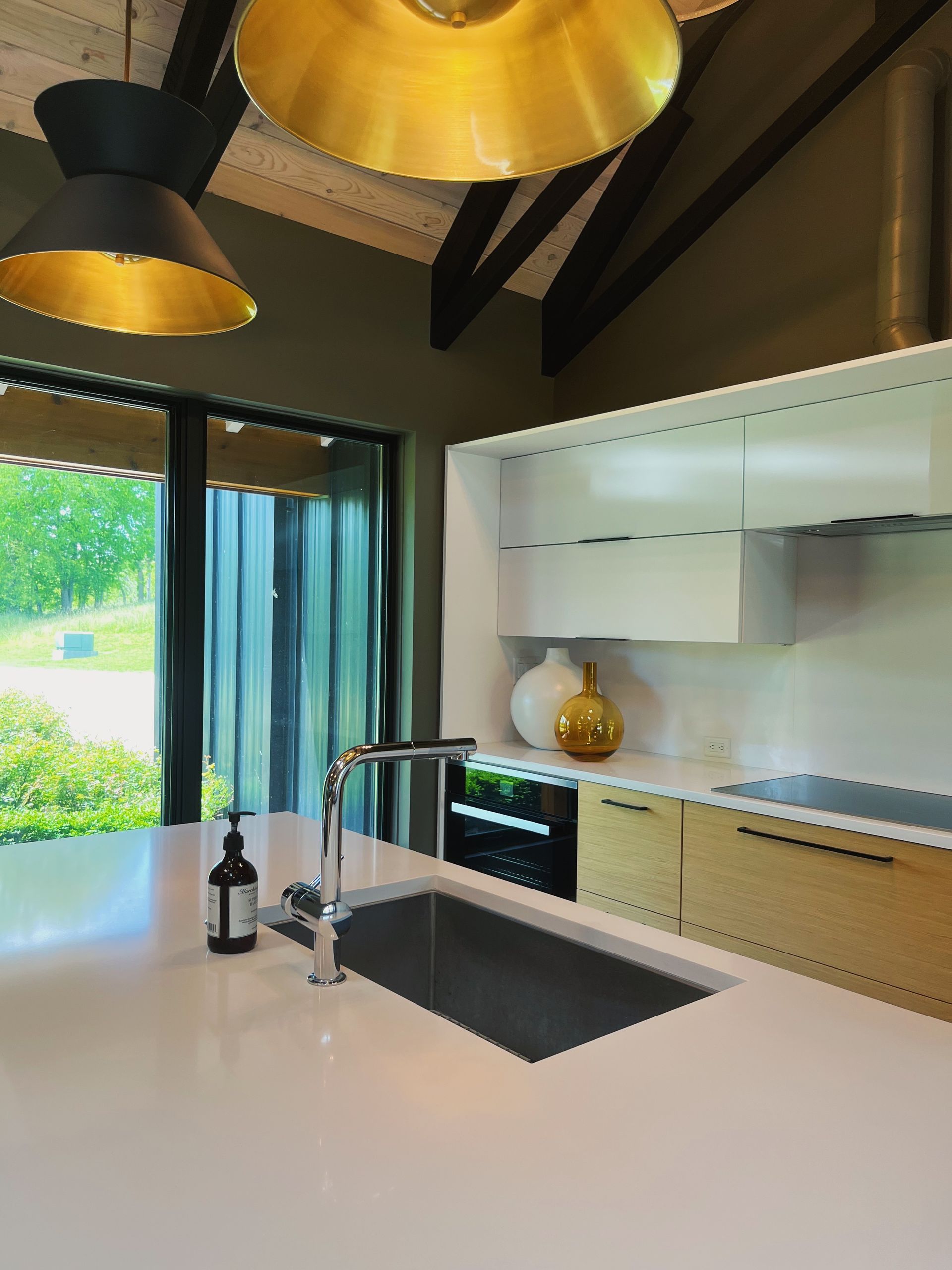 Modern kitchen with white island, black sink, brass pendant lights, and sliding glass doors to greenery
