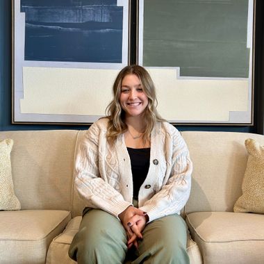 Smiling woman seated on a cream sofa in front of abstract wall art, wearing a white cardigan and green pants.