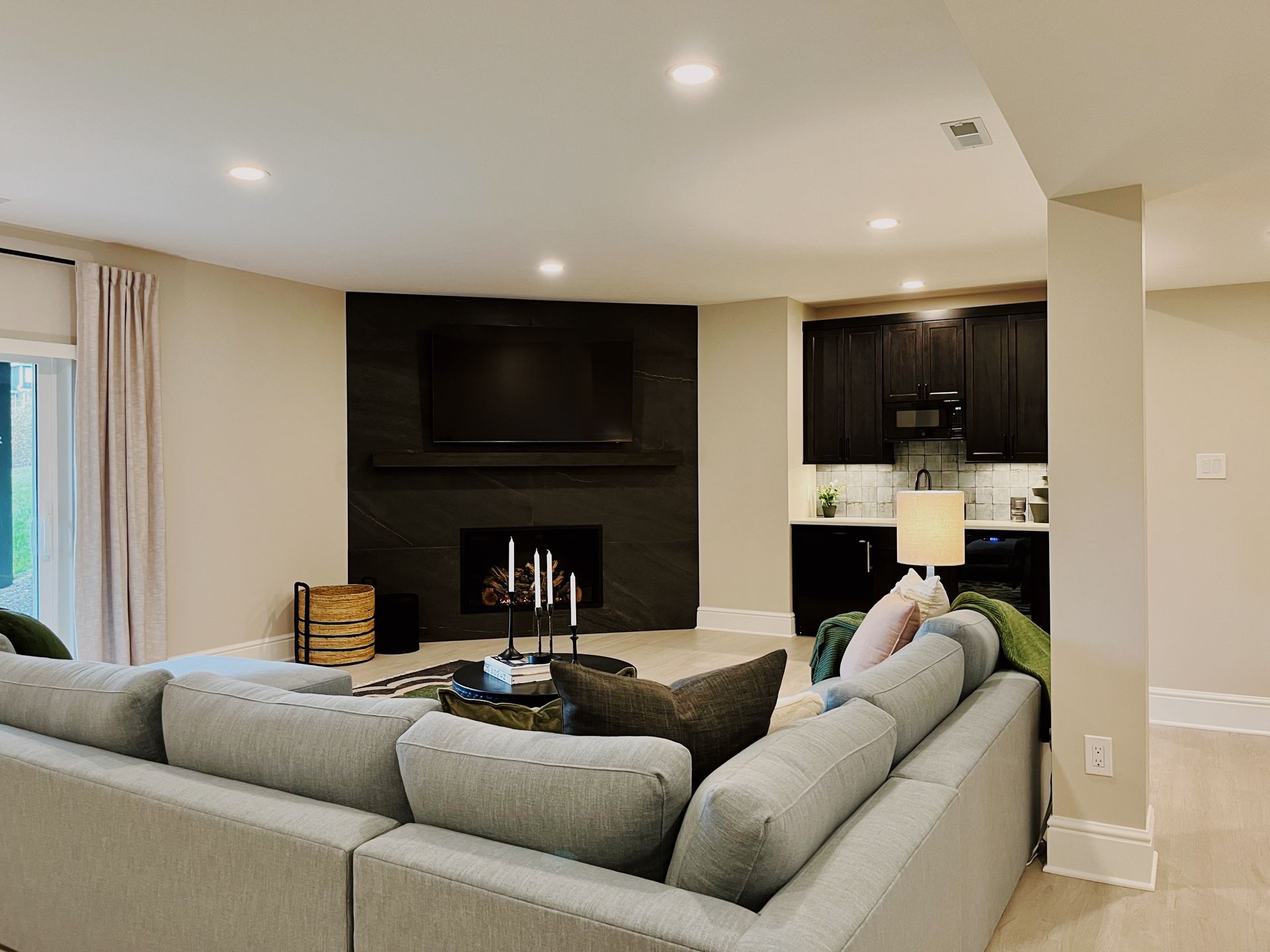 Bright living room with gray sectional, fireplace, wall-mounted TV, and dark kitchen cabinets.
