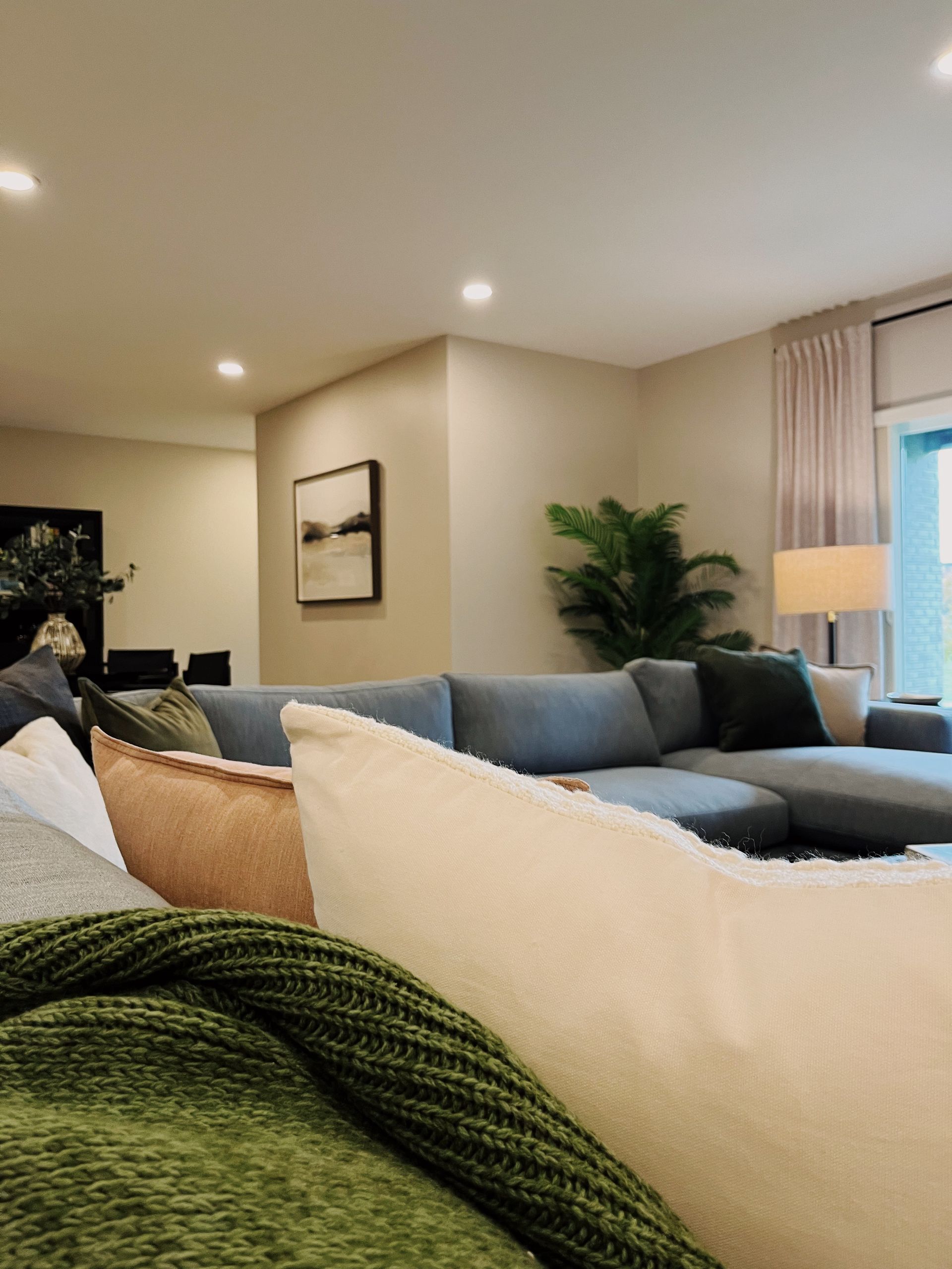 Bright living room with a sectional sofa, cream pillows, green throw, recessed lights, and a window with curtains.