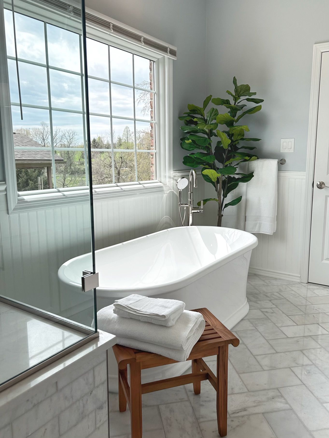 Bright bathroom with freestanding tub, walk-in shower, wood stool, and large window.