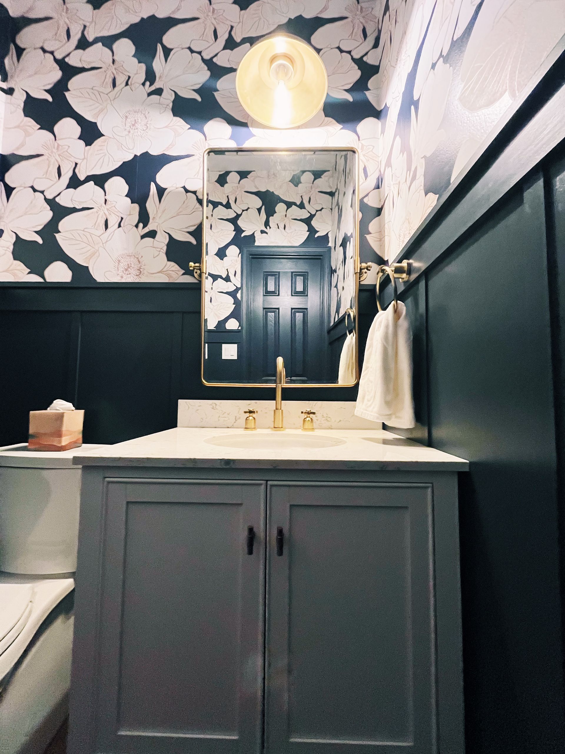 Bathroom vanity with dark floral wallpaper, black wainscoting, white sink, and round lighted mirror