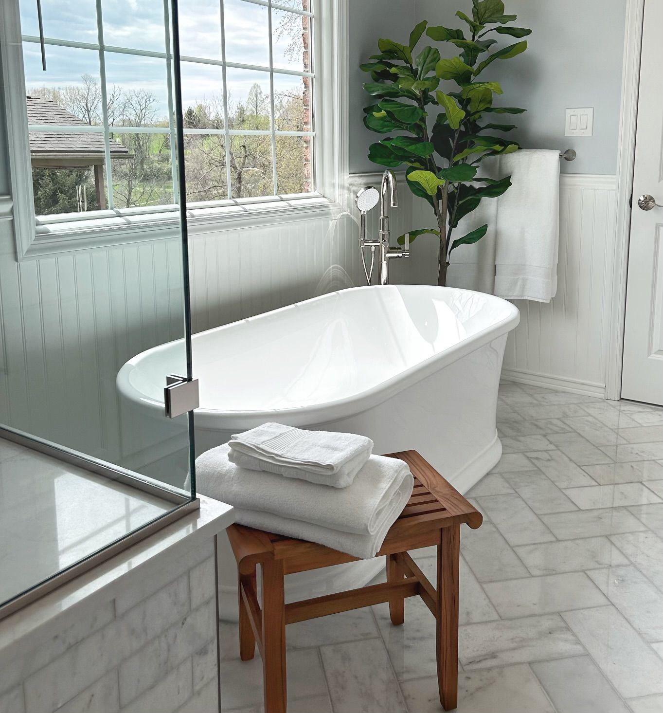 Bright bathroom with white freestanding tub, wood stool, towels, large window, and potted plant.