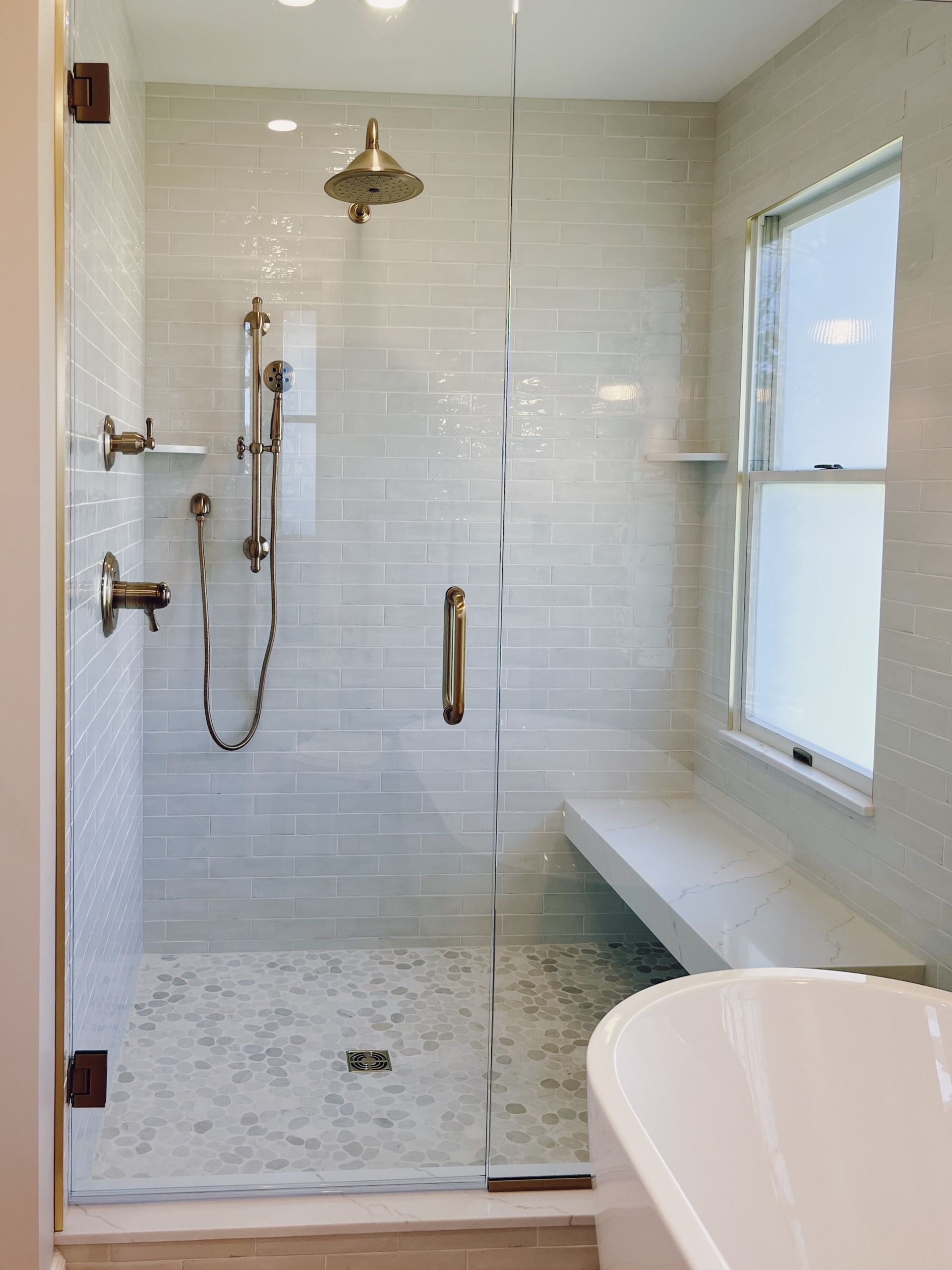 Modern shower with glass door, white tile, brass fixtures, and a window bench