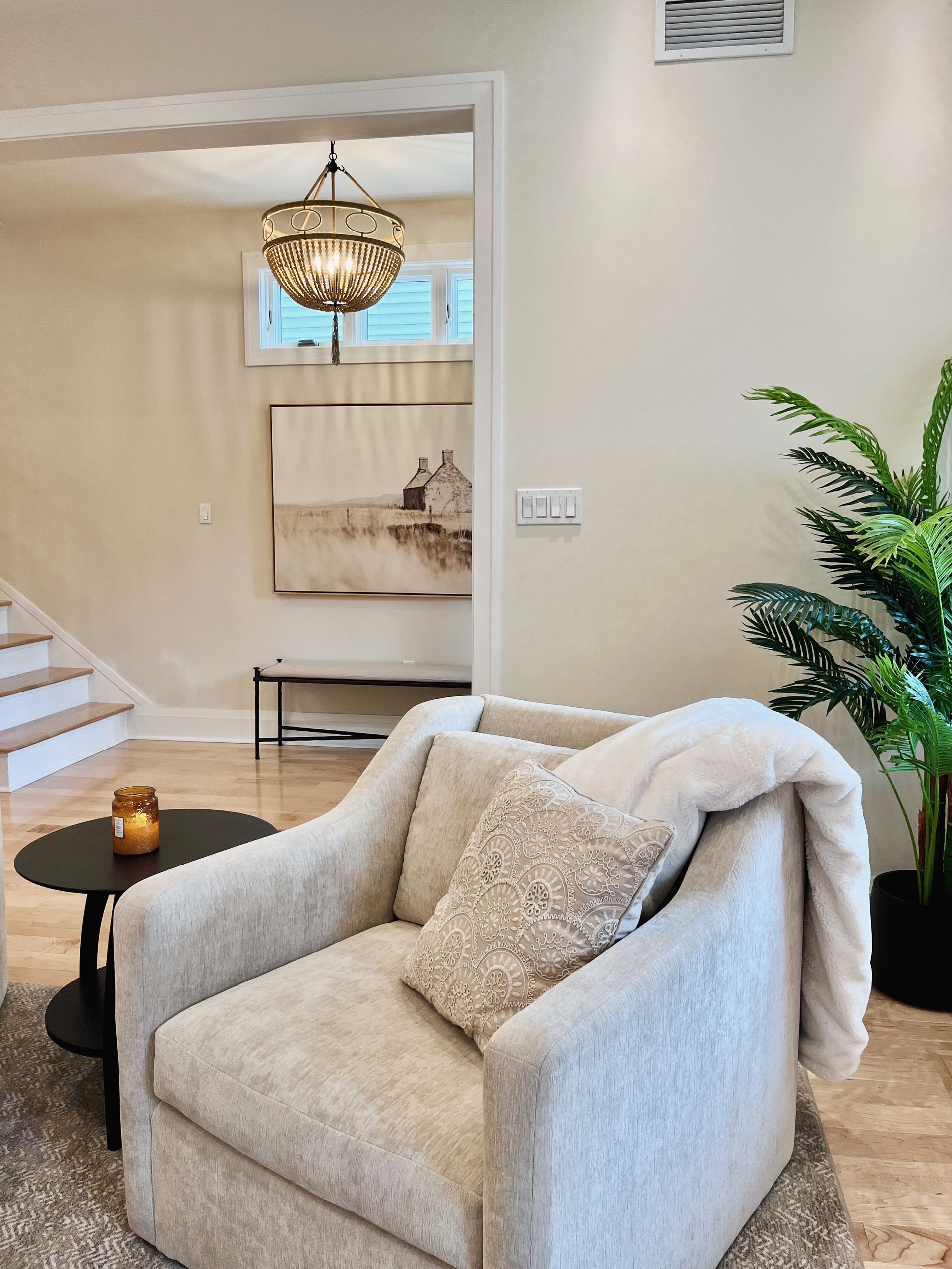 Bright living room with a beige armchair, black side table, chandelier, and potted plant near a staircase.
