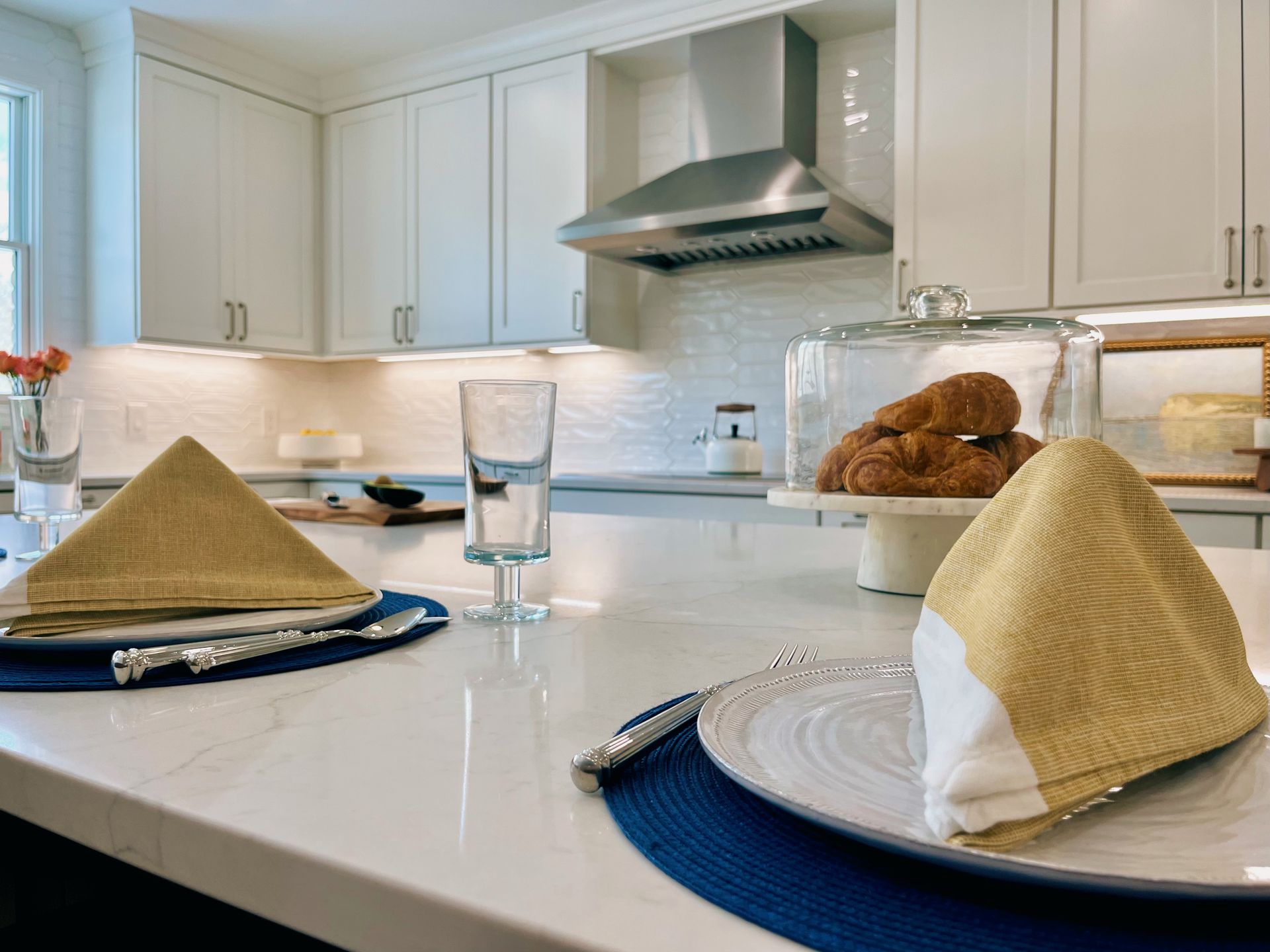 Modern white kitchen with set dining table, blue placemats, and covered plates in the foreground