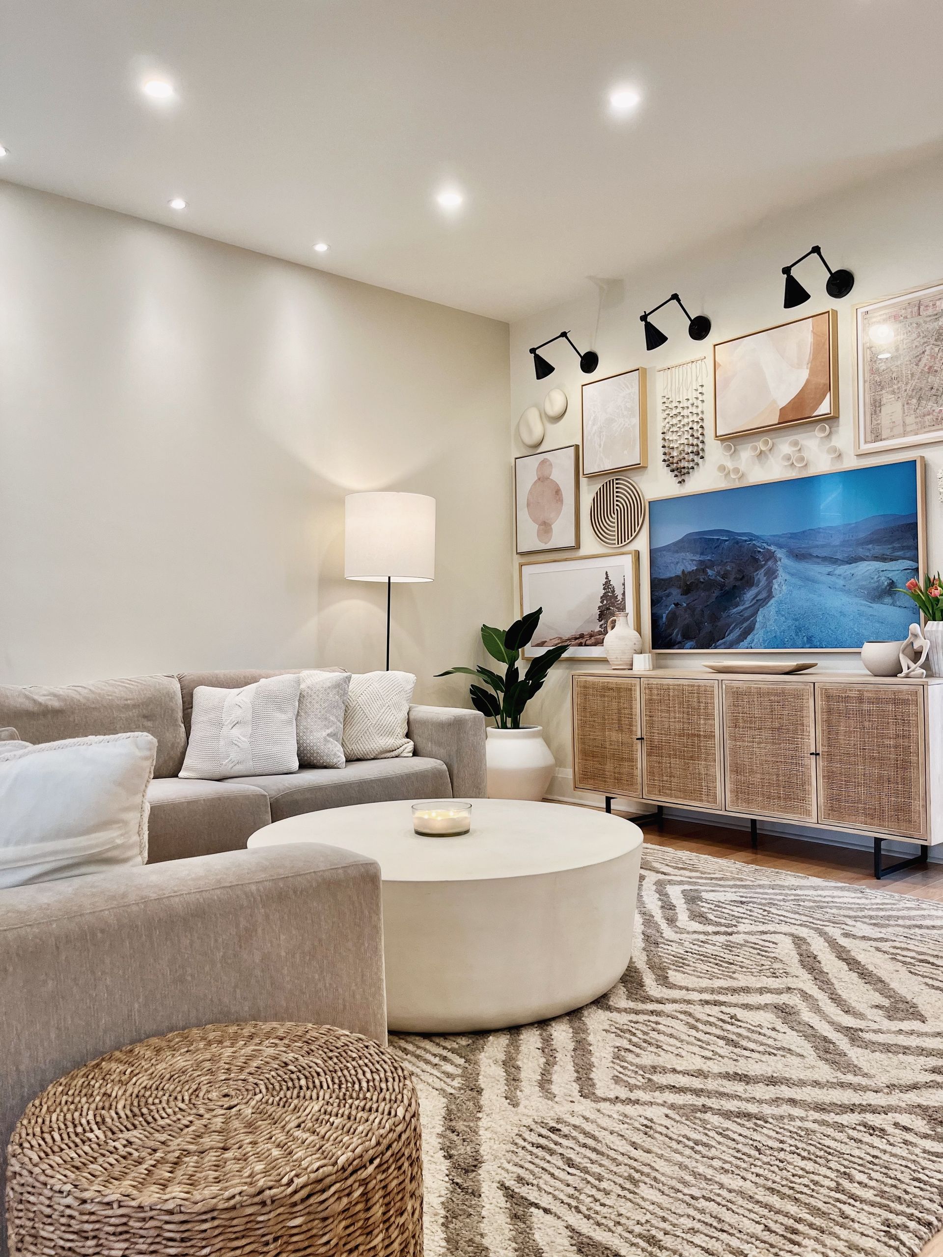 Modern living room with neutral-toned furniture, round white coffee table, woven stool, and large framed wall art.