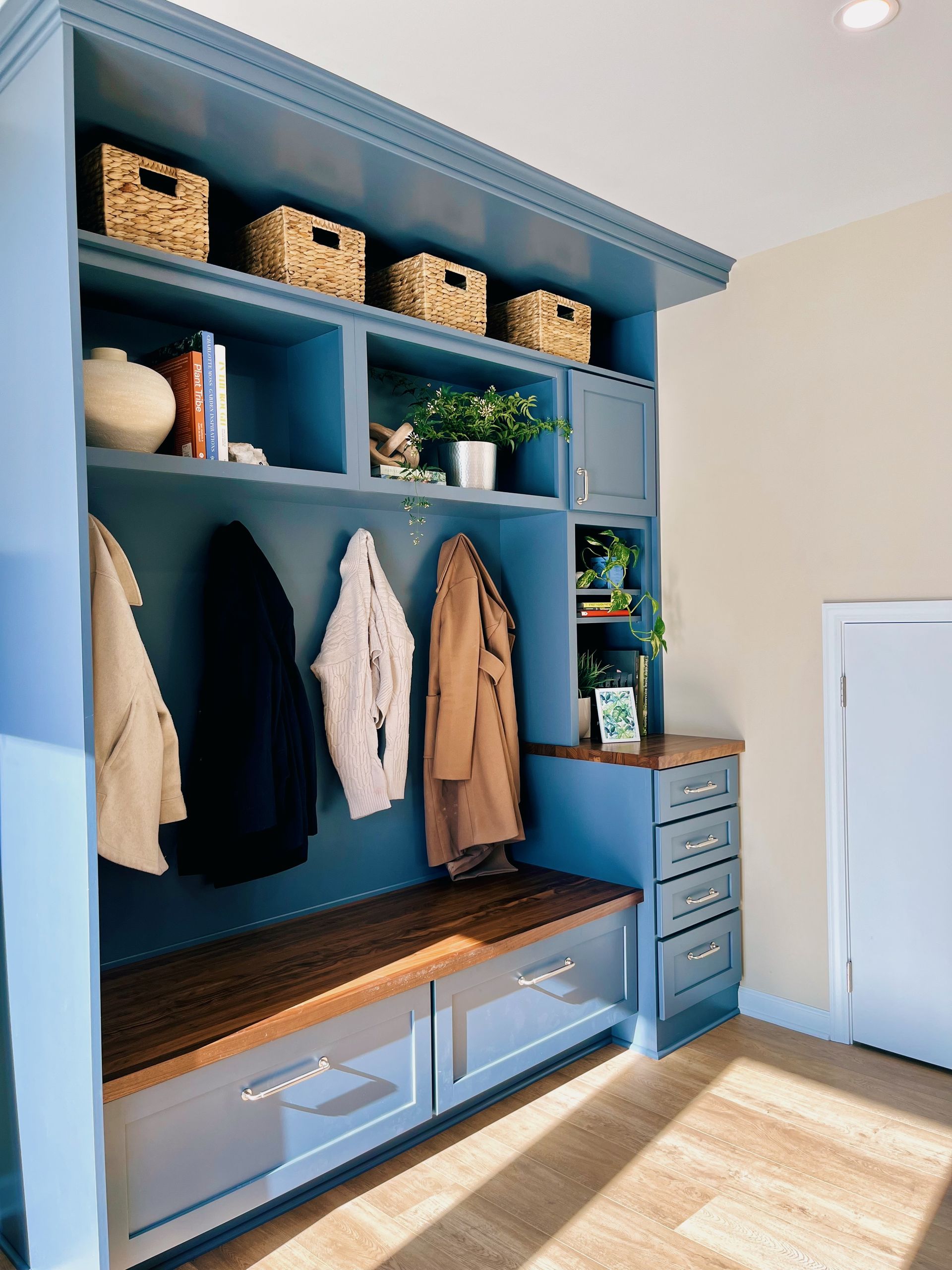 Blue mudroom with built-in cubbies, hanging coats, storage baskets, and drawers in bright sunlight