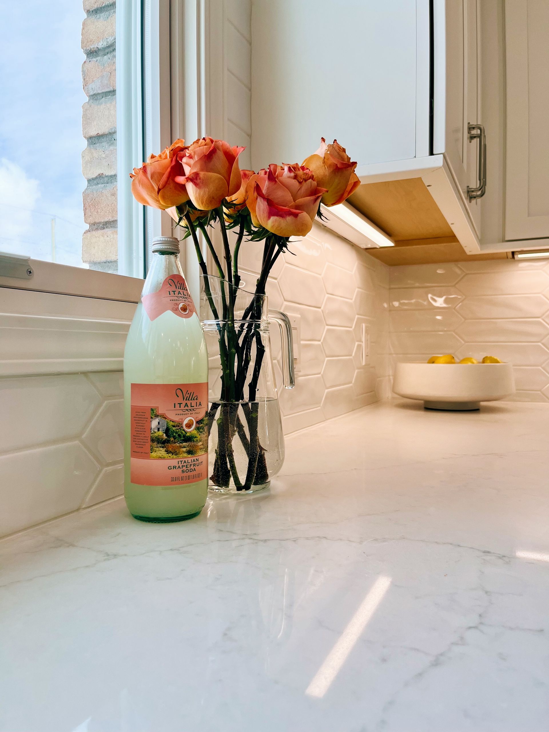 Pink flowers in a glass vase beside a pastel bottle on a white kitchen countertop