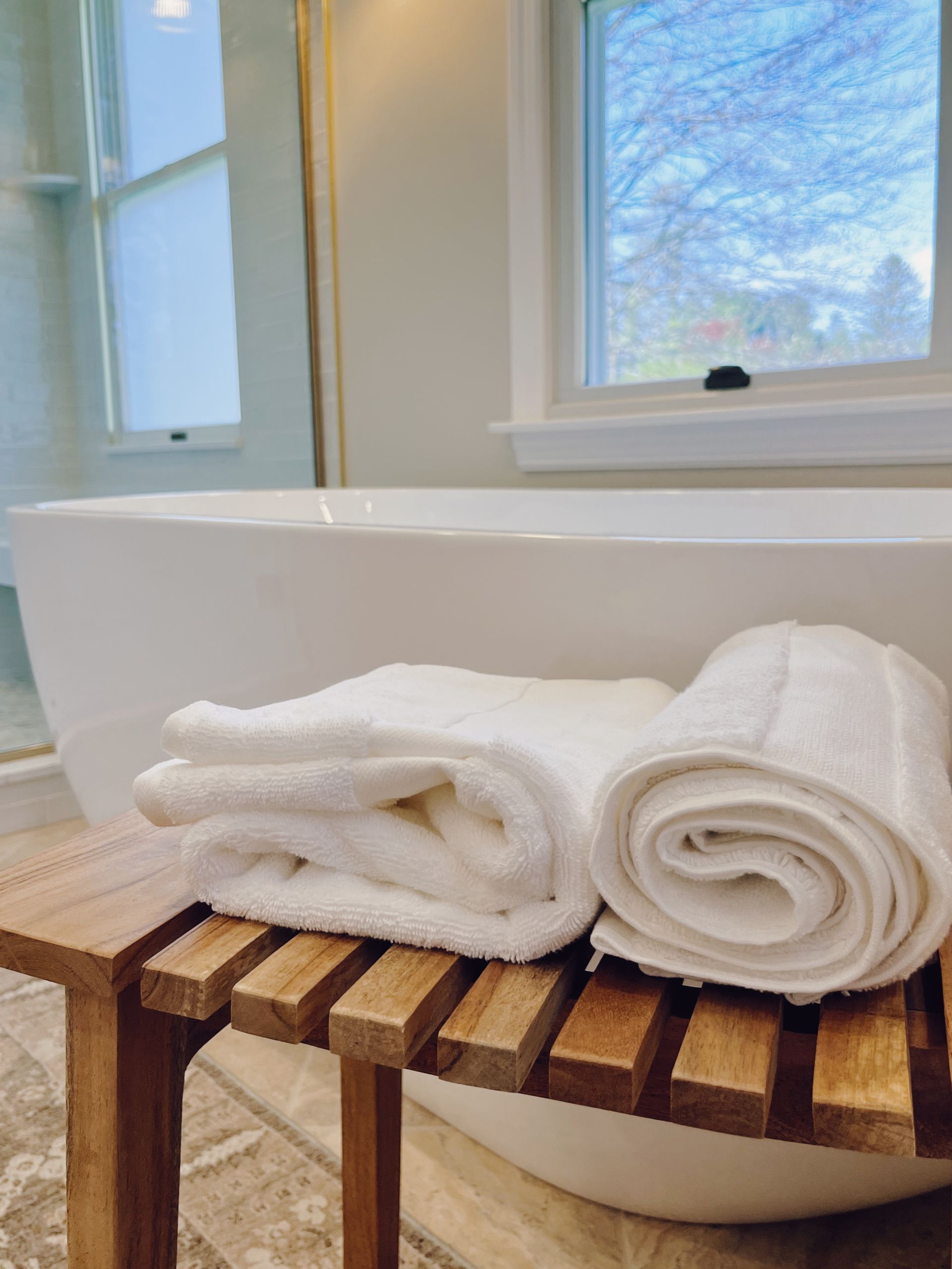 White bathroom with freestanding tub and wooden bench holding rolled white towels