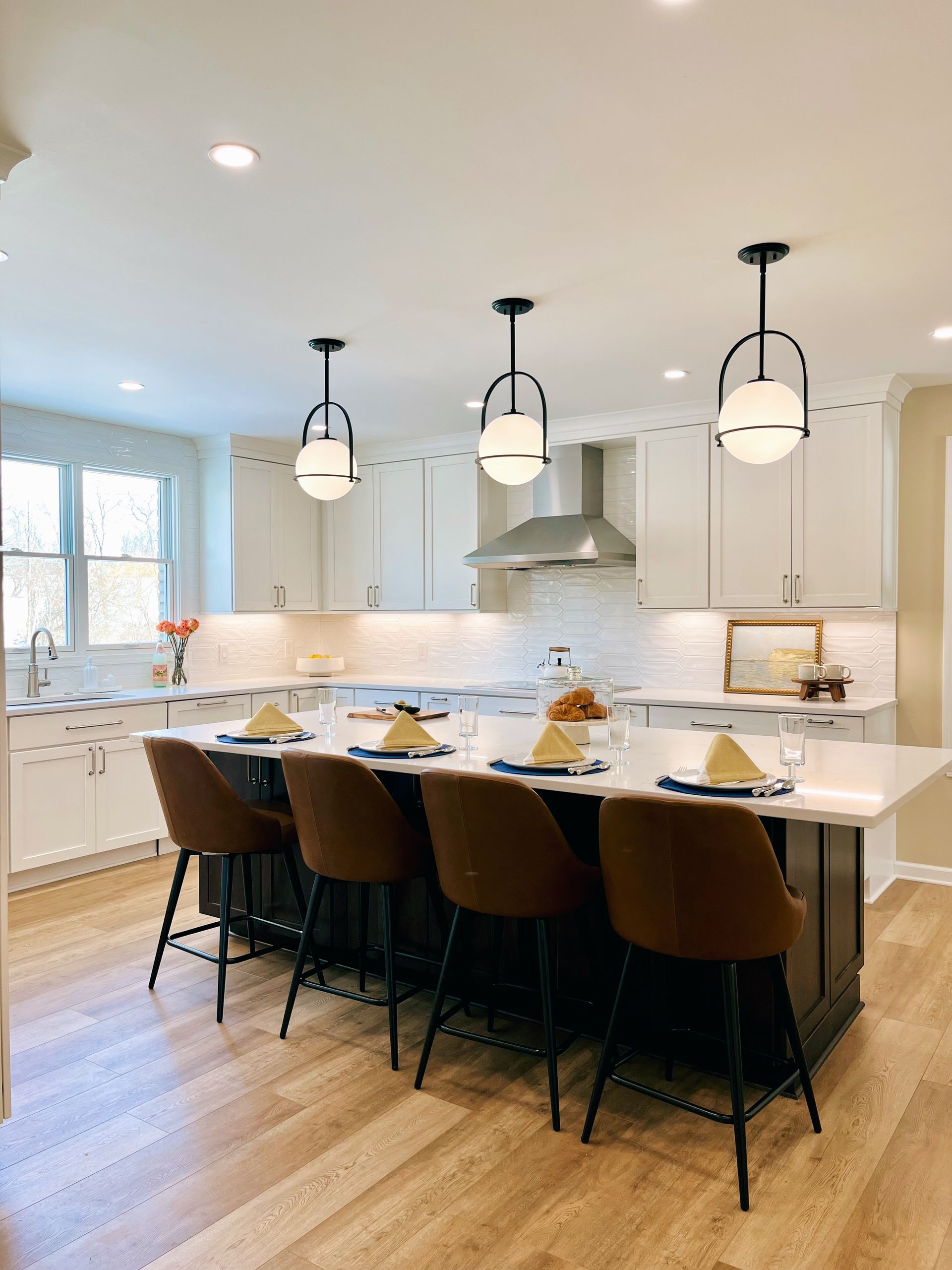 Modern white kitchen with a large island, black stools, pendant lights, and hardwood floors