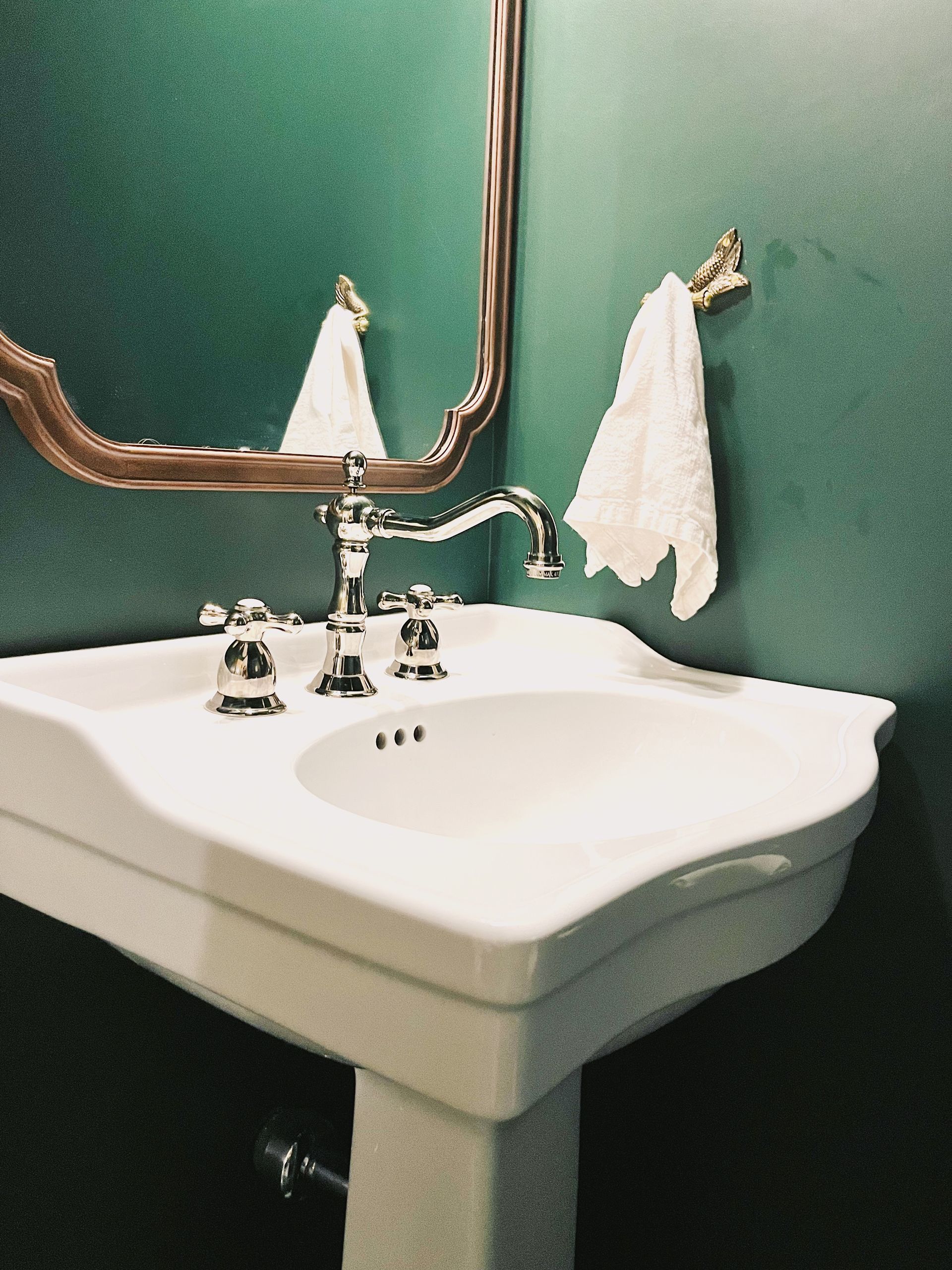 White bathroom sink with brass faucet and mirror against a green wall, with two hanging hand towels