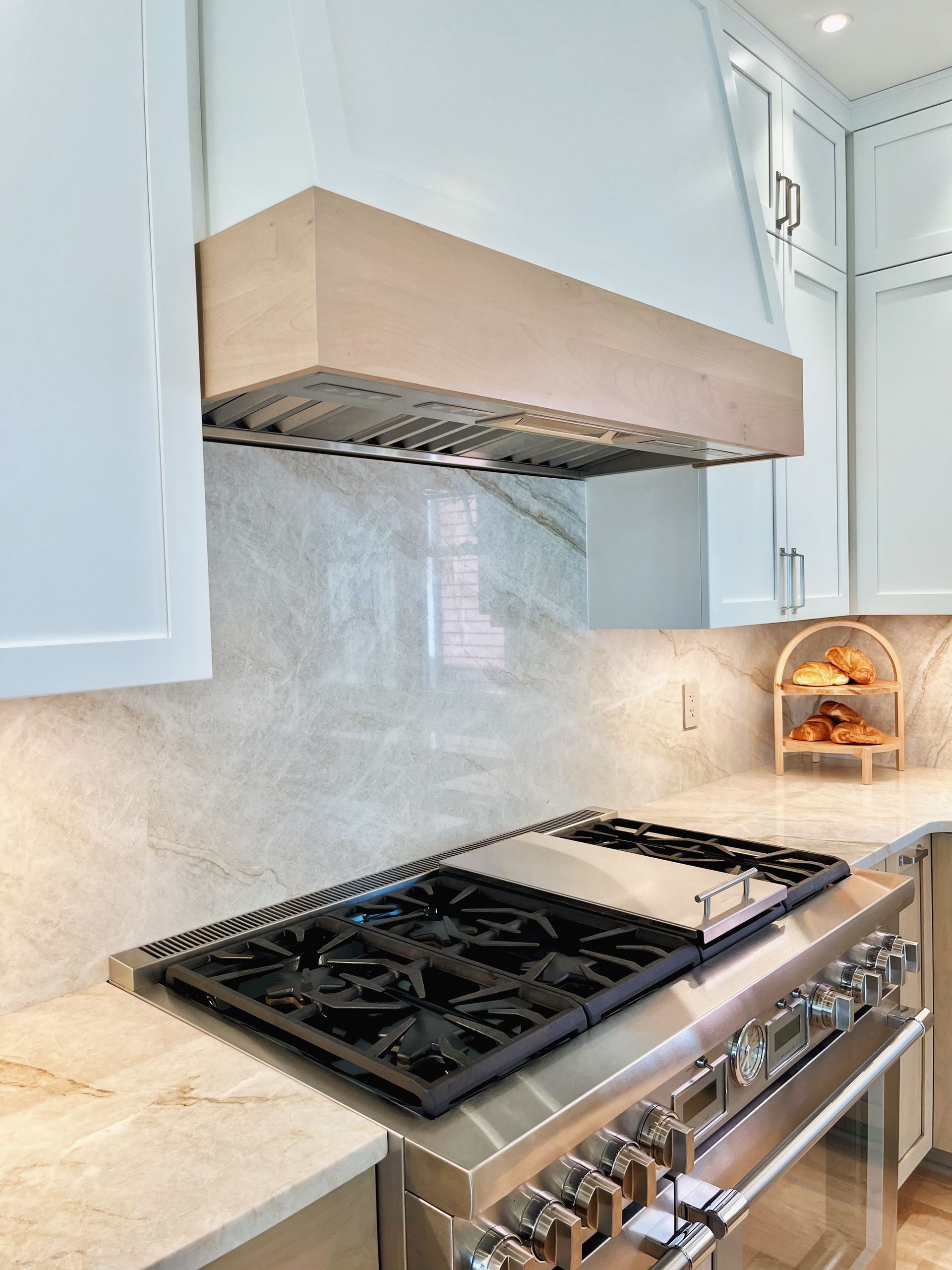 Modern kitchen with stainless gas cooktop, light wood range hood, white cabinets, and marble backsplash