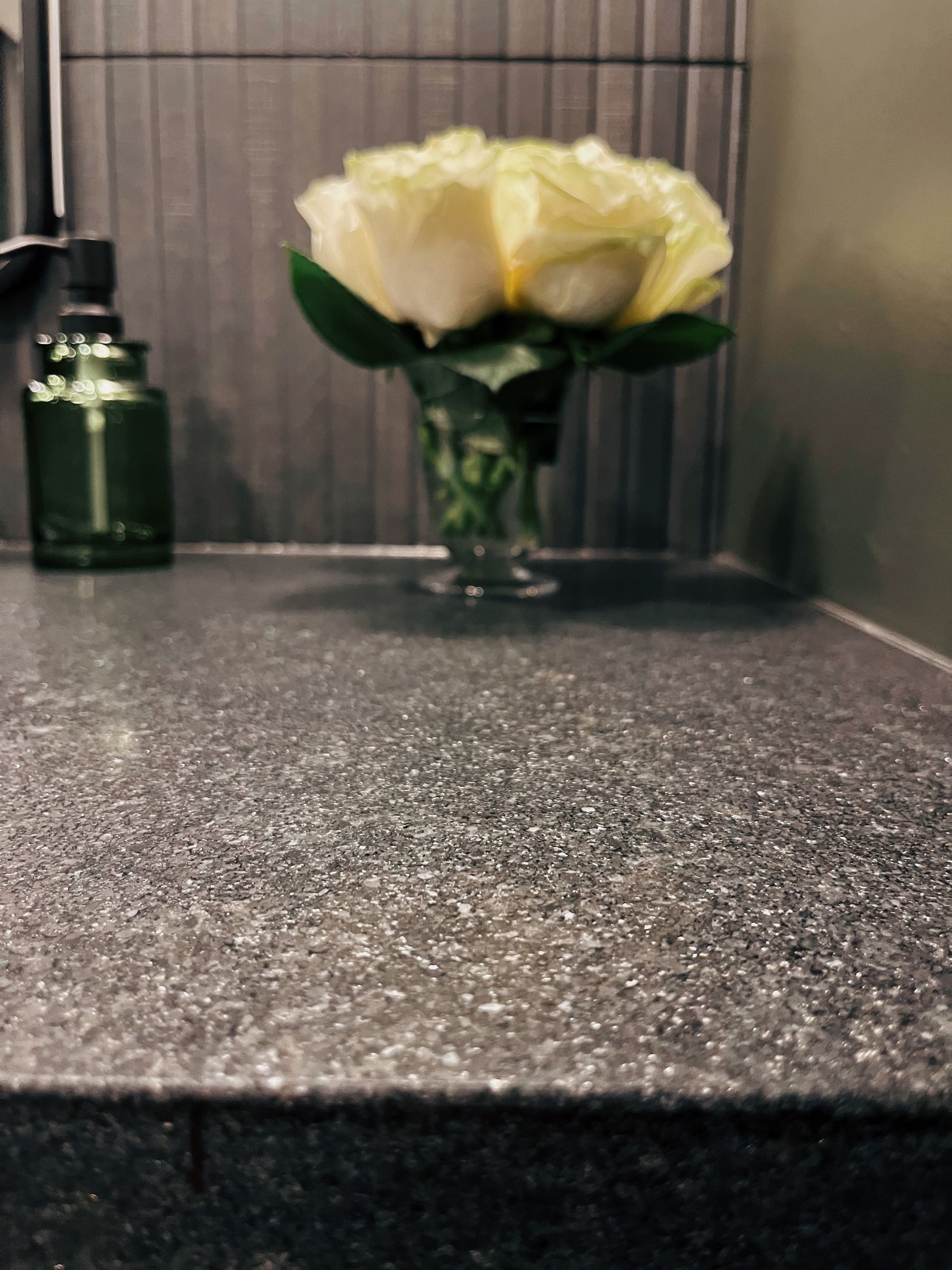Yellow flowers in a glass vase on a dark speckled countertop beside a green bottle.