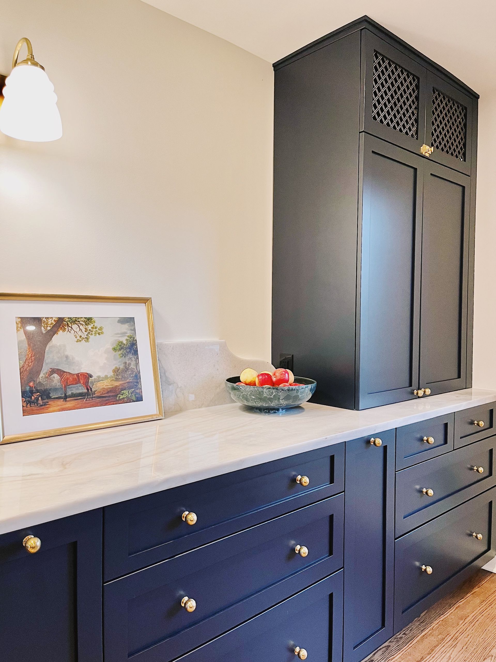 Blue kitchen cabinets with gold knobs, white countertop, framed art, fruit bowl, and tall dark pantry cabinet