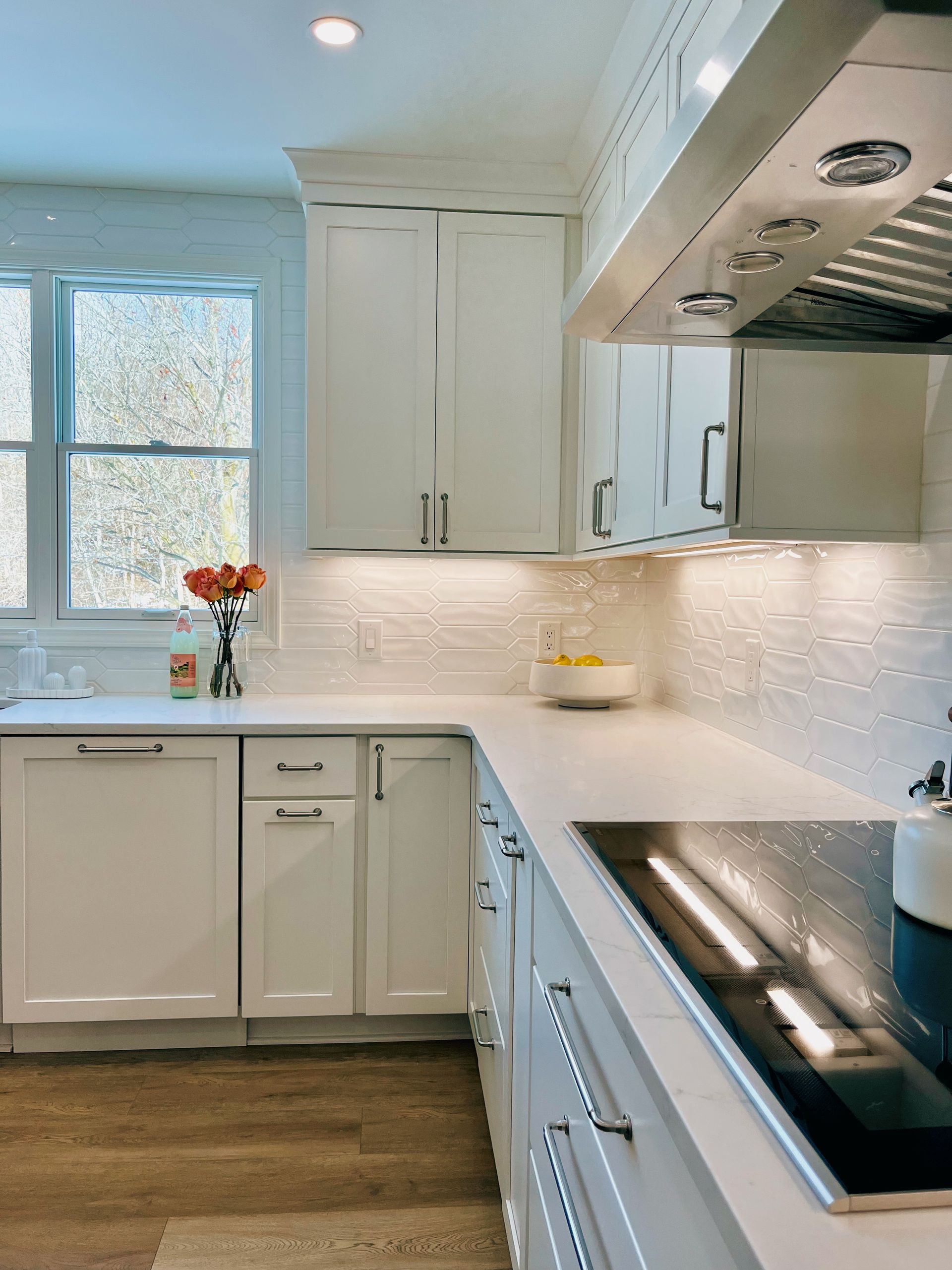 Bright modern white kitchen with marble backsplash, wood floor, and black cooktop by a window.