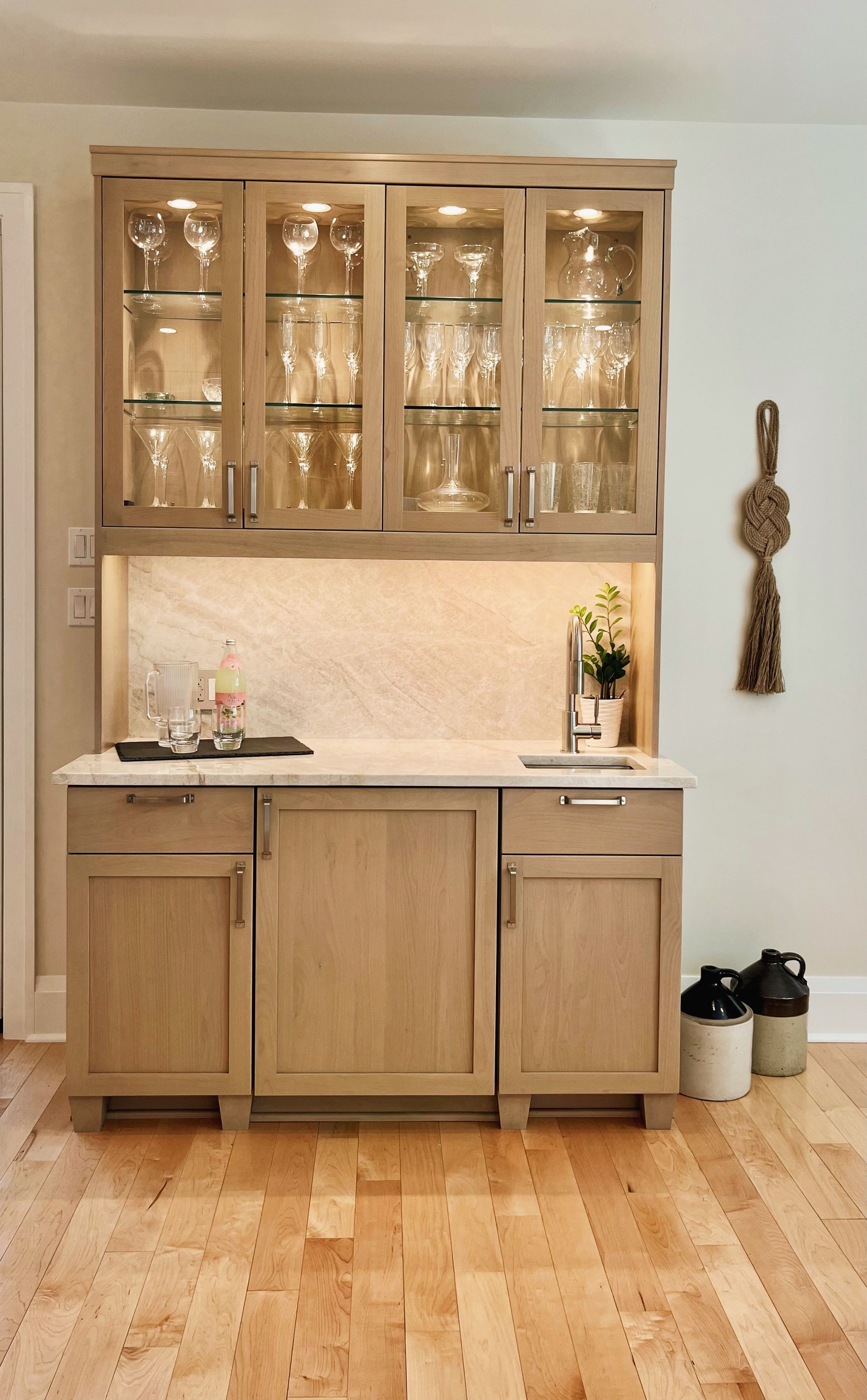 Wooden bar cabinet with glass doors and countertop in a light-colored room, with a floor broom and shoes nearby