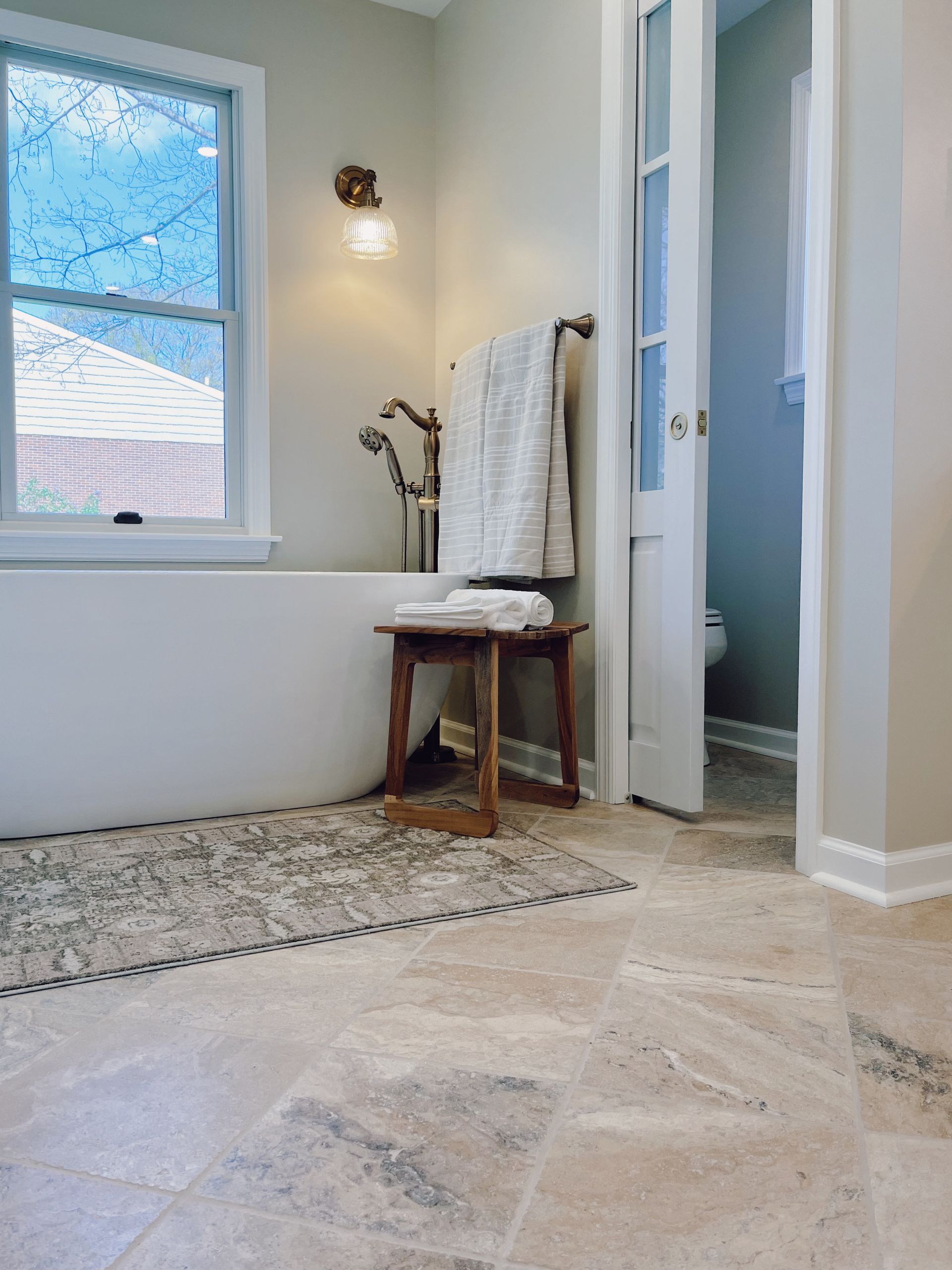 Bright bathroom with a freestanding tub, window, towel rack, and light tile floor.