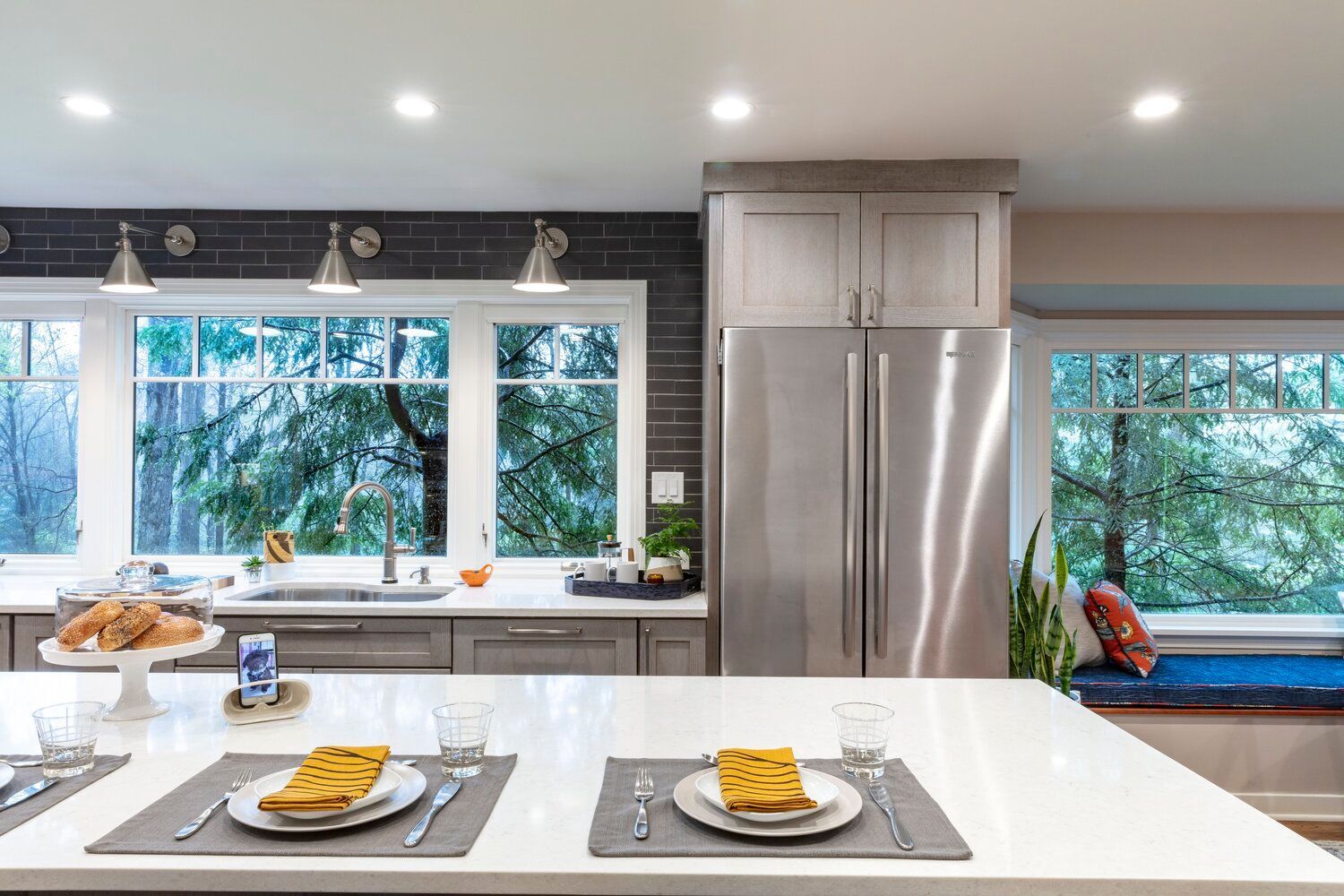 Modern kitchen with white island, place settings, stainless fridge, and large windows overlooking greenery