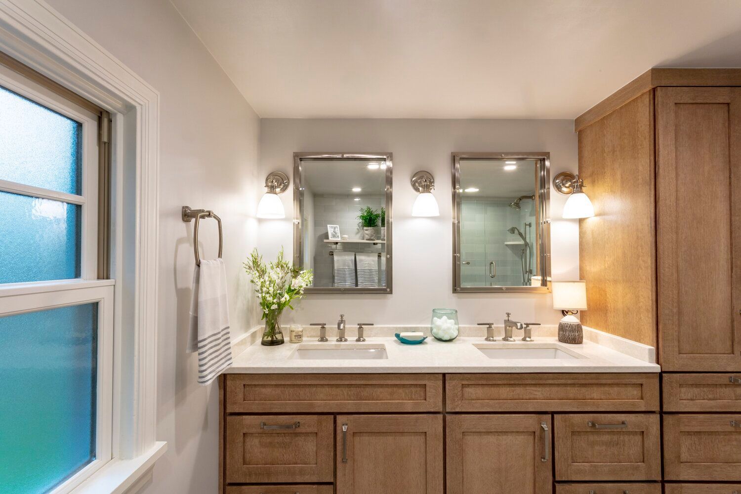 Bright bathroom vanity with two mirrors, wooden cabinets, white countertop, and frosted window.