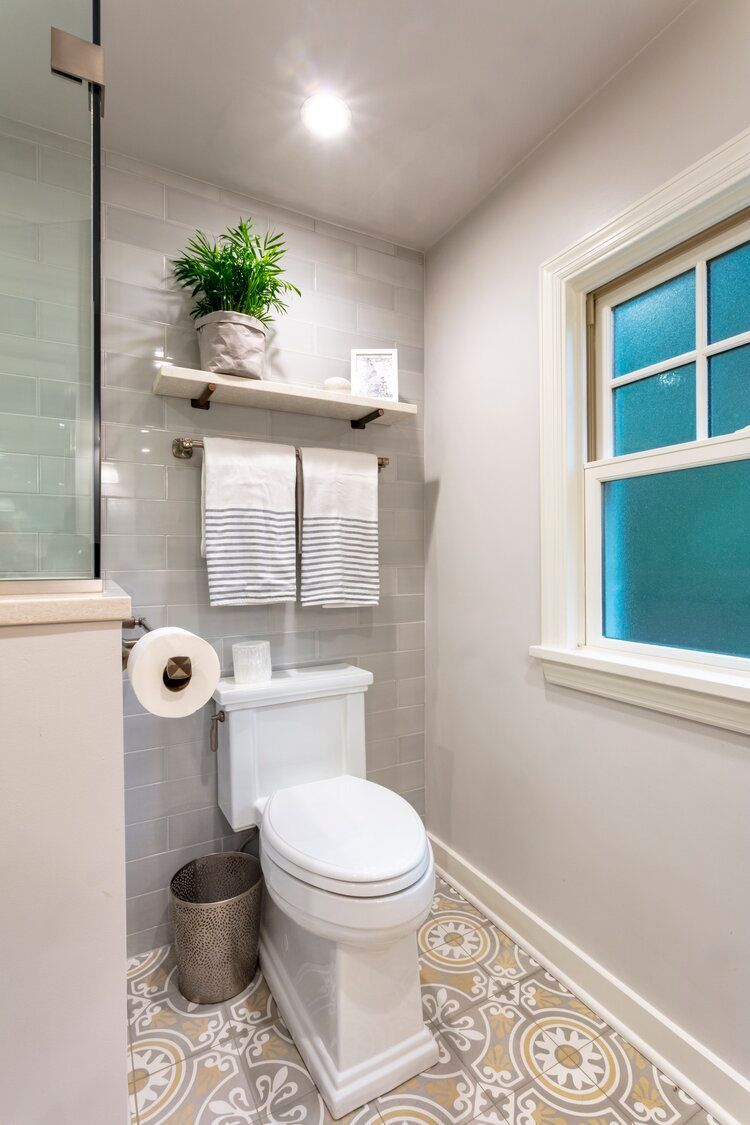 Small bathroom with toilet, patterned tile floor, floating shelf, and window with blue blind.
