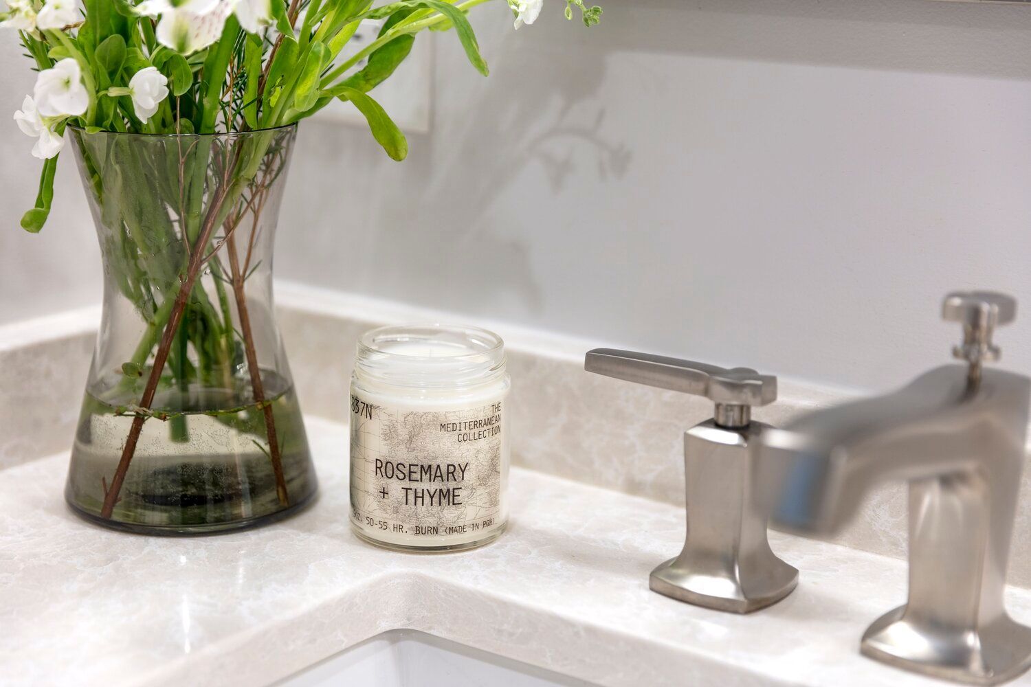 White bathroom sink with a vase of white flowers, a candle, and chrome faucet on the countertop