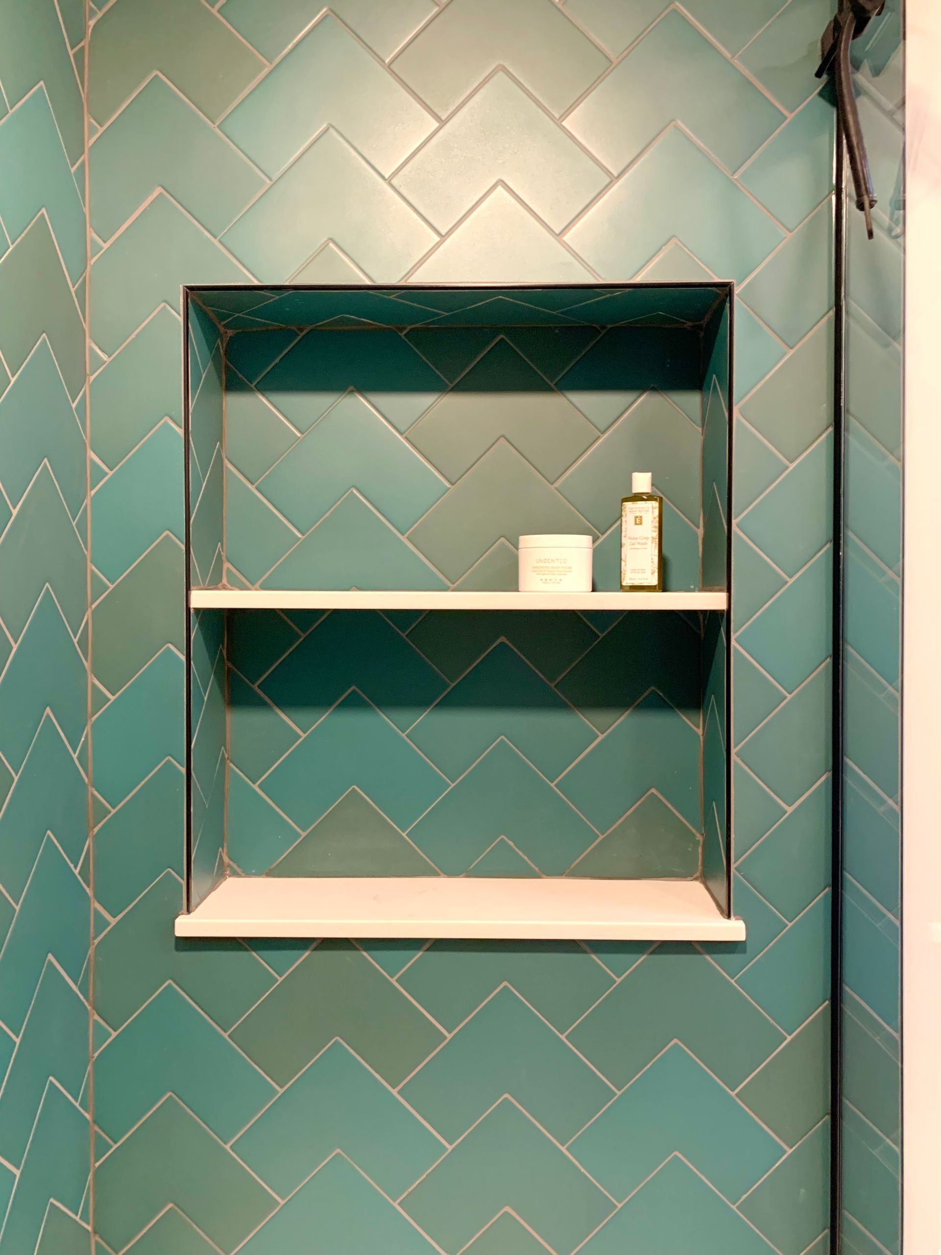 Bathroom niche with turquoise herringbone tile and three shelves holding small toiletries.
