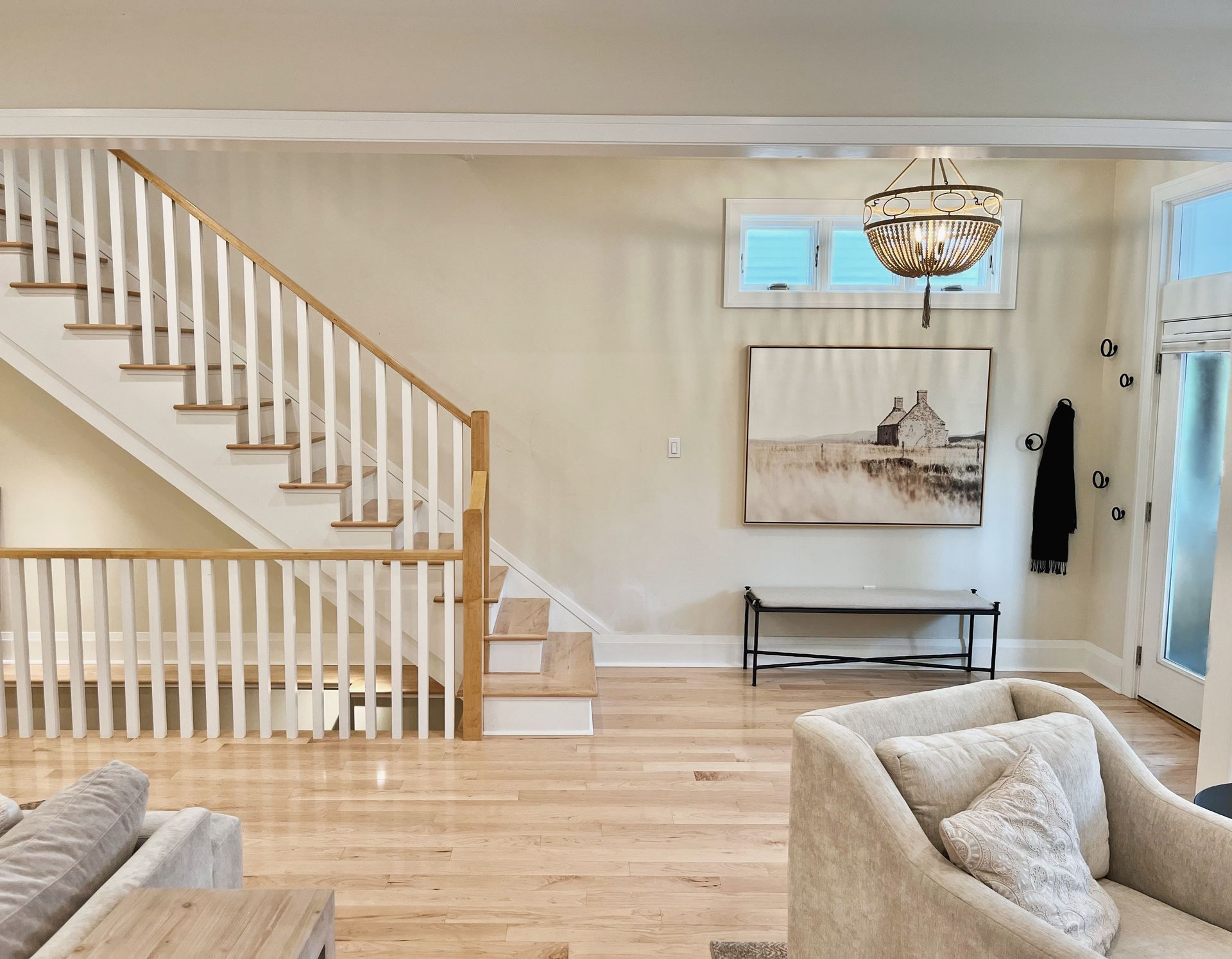 Bright living room with hardwood floors, staircase, beige sofa, wall art, and a chandelier.