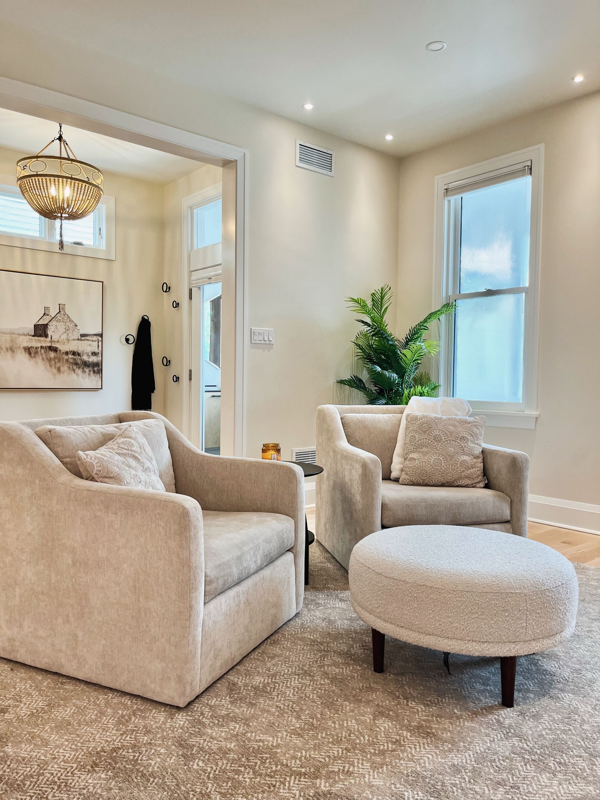 Bright living room with beige chairs, ottoman, plant, and large windows