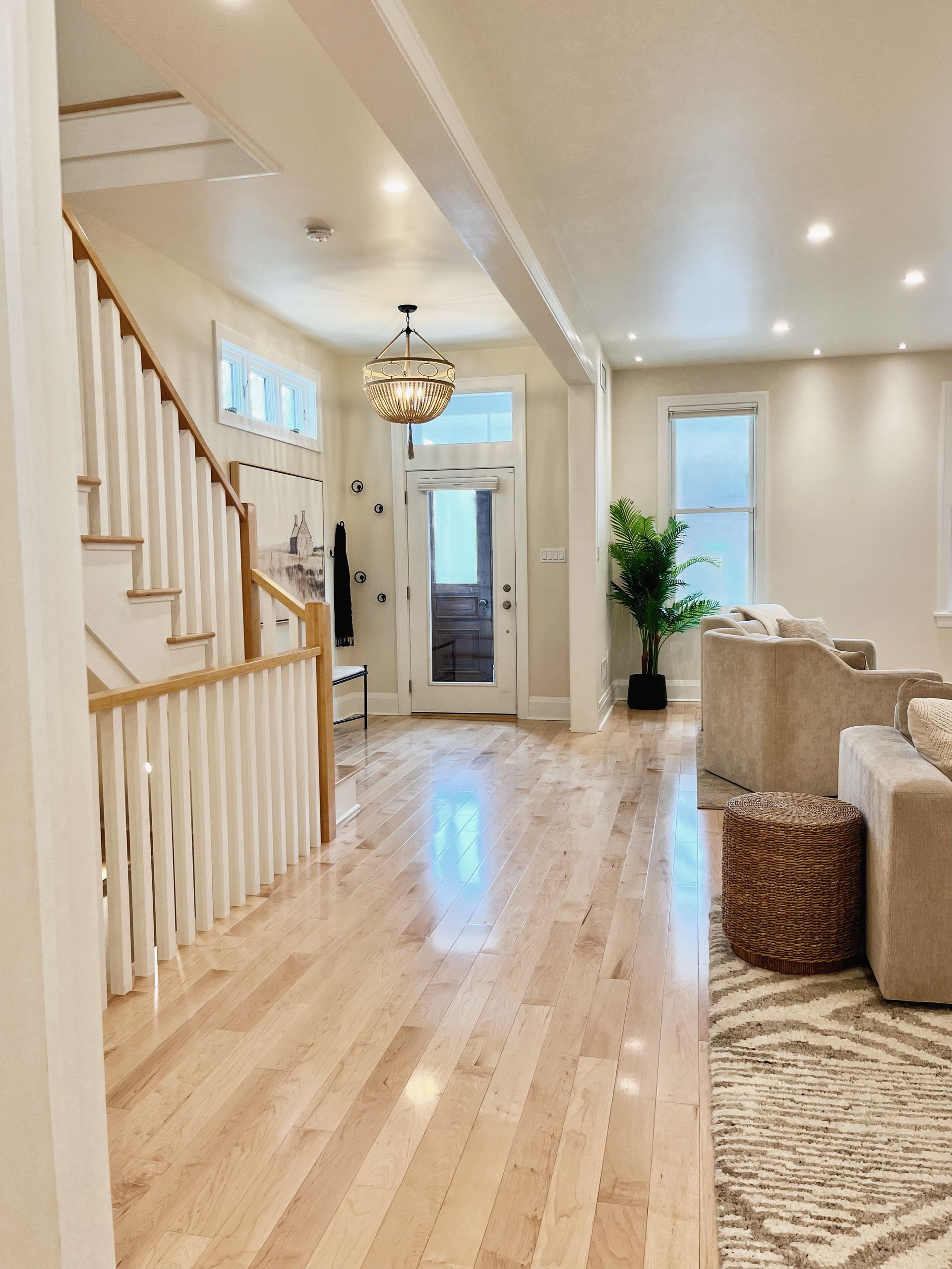 Bright foyer with staircase, hardwood floors, front door, hanging light, and beige couch seating area