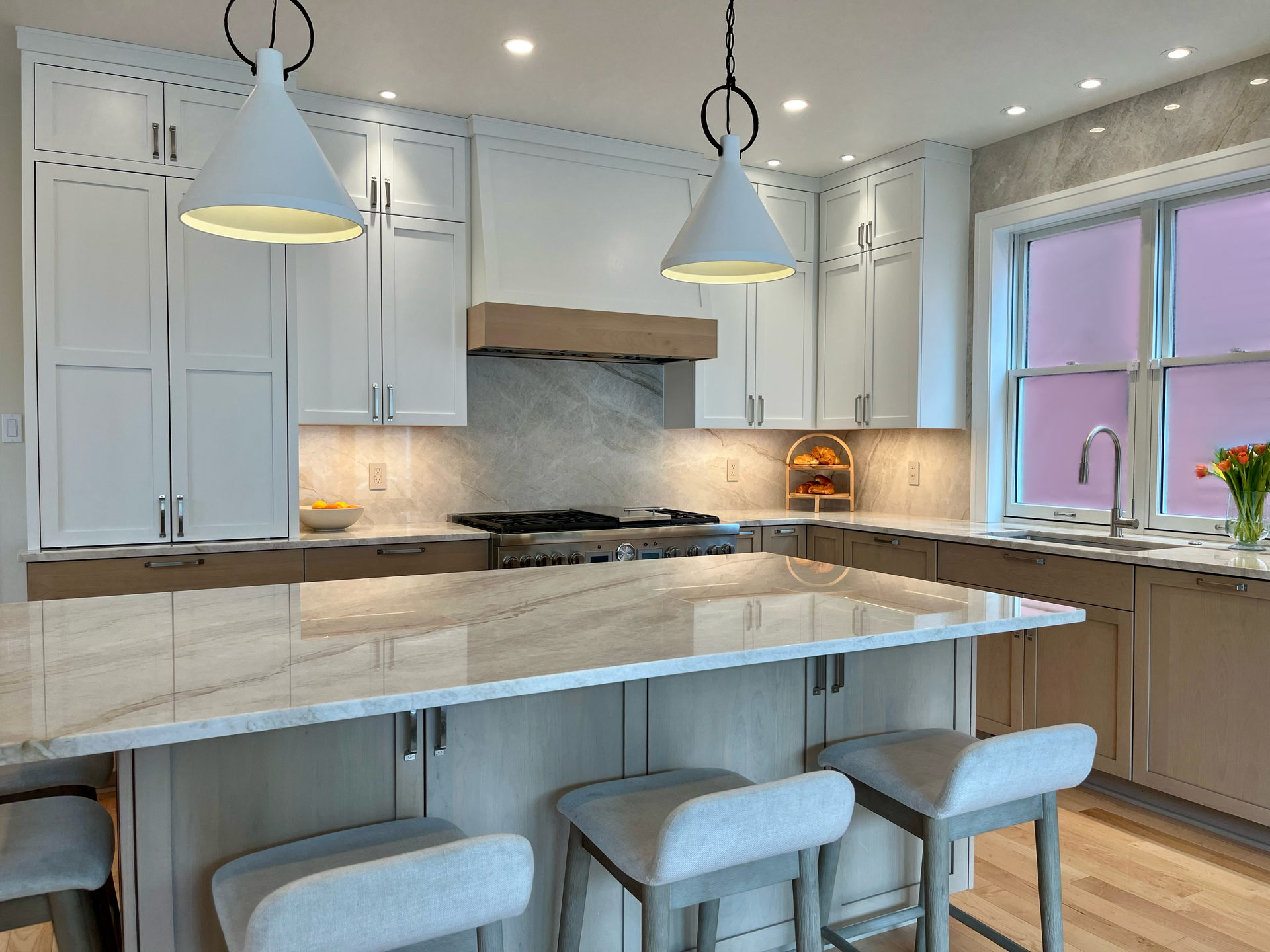 Modern white kitchen with marble island, pendant lights, bar stools, and a window over the sink.