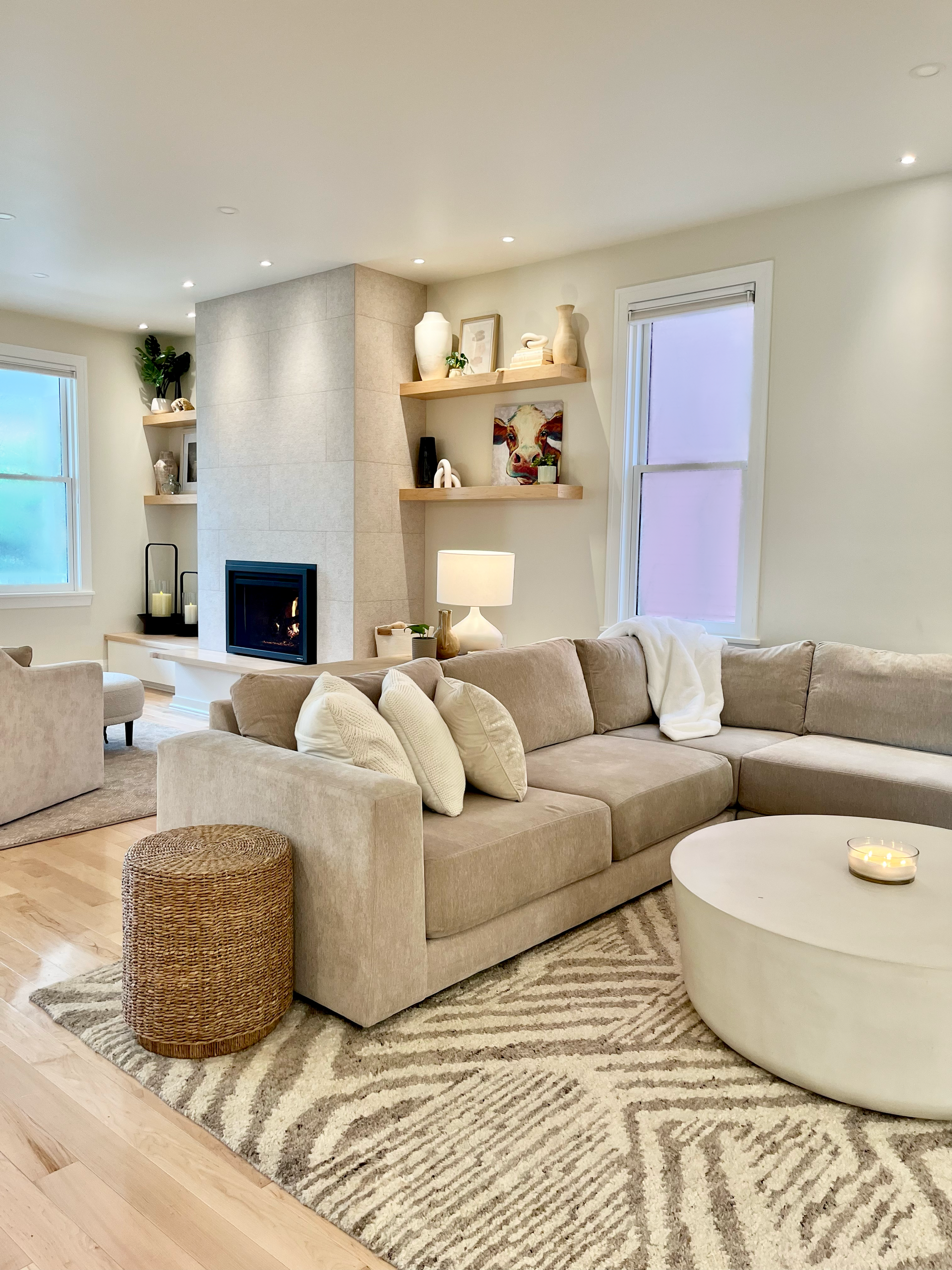 Bright beige living room with sectional sofa, round ottoman, woven pouf, fireplace, and built-in shelves