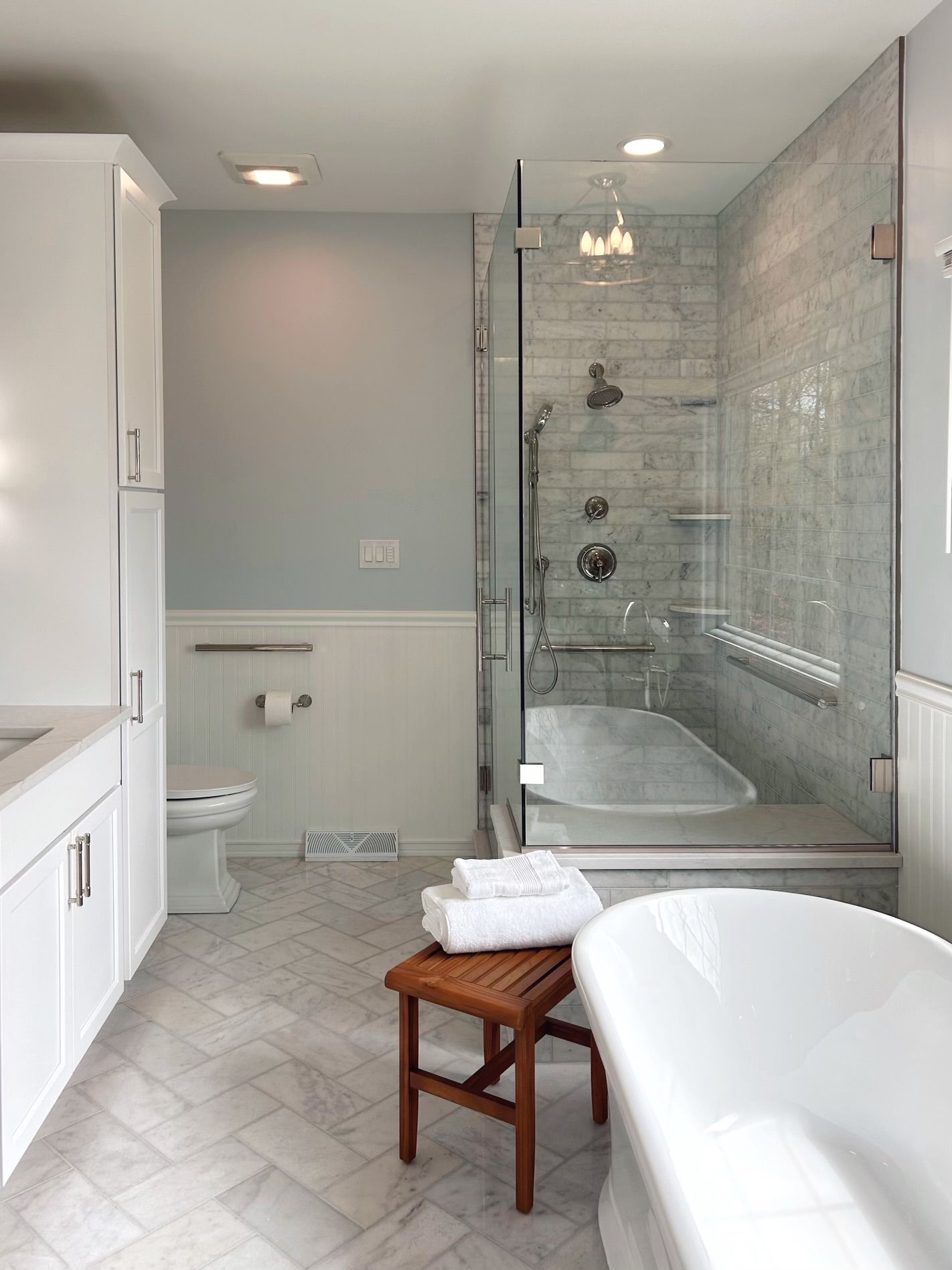 Bright modern bathroom with glass shower, bathtub, toilet, and a wooden stool with folded towels.