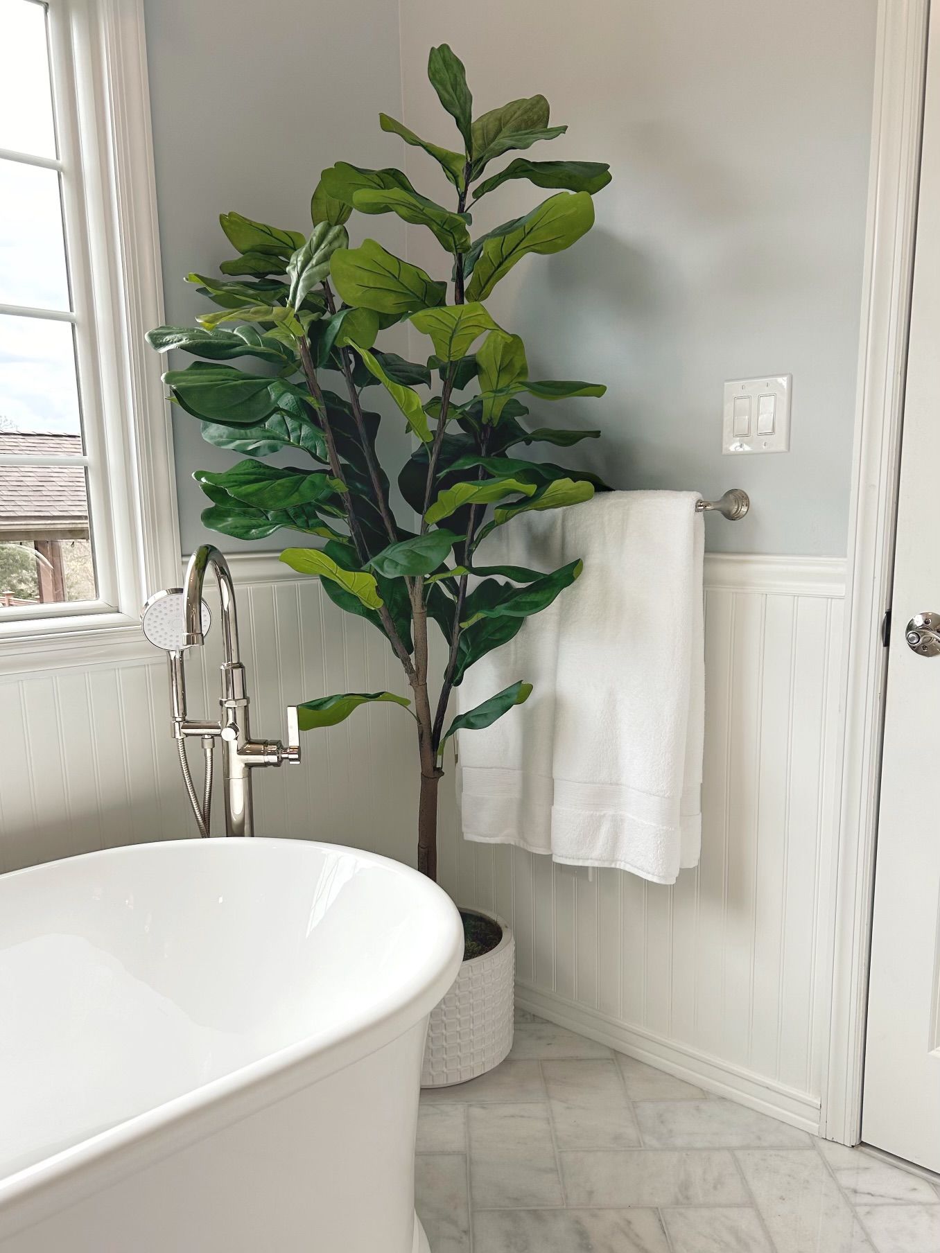 Bright bathroom with a white tub, potted plant, towel rack, and marble-style floor.
