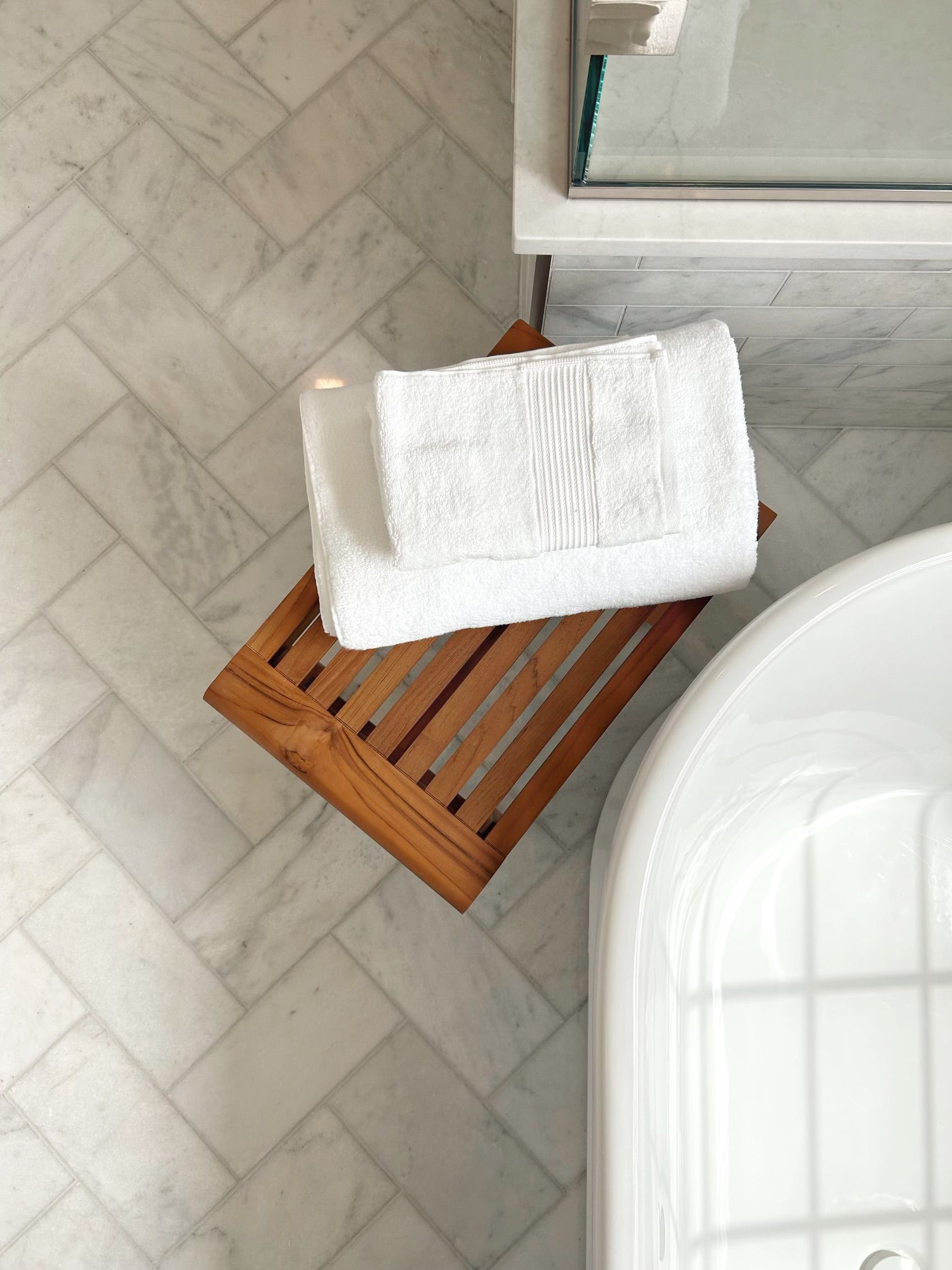 White towel on a wooden stool beside a white bathtub on a tiled bathroom floor.