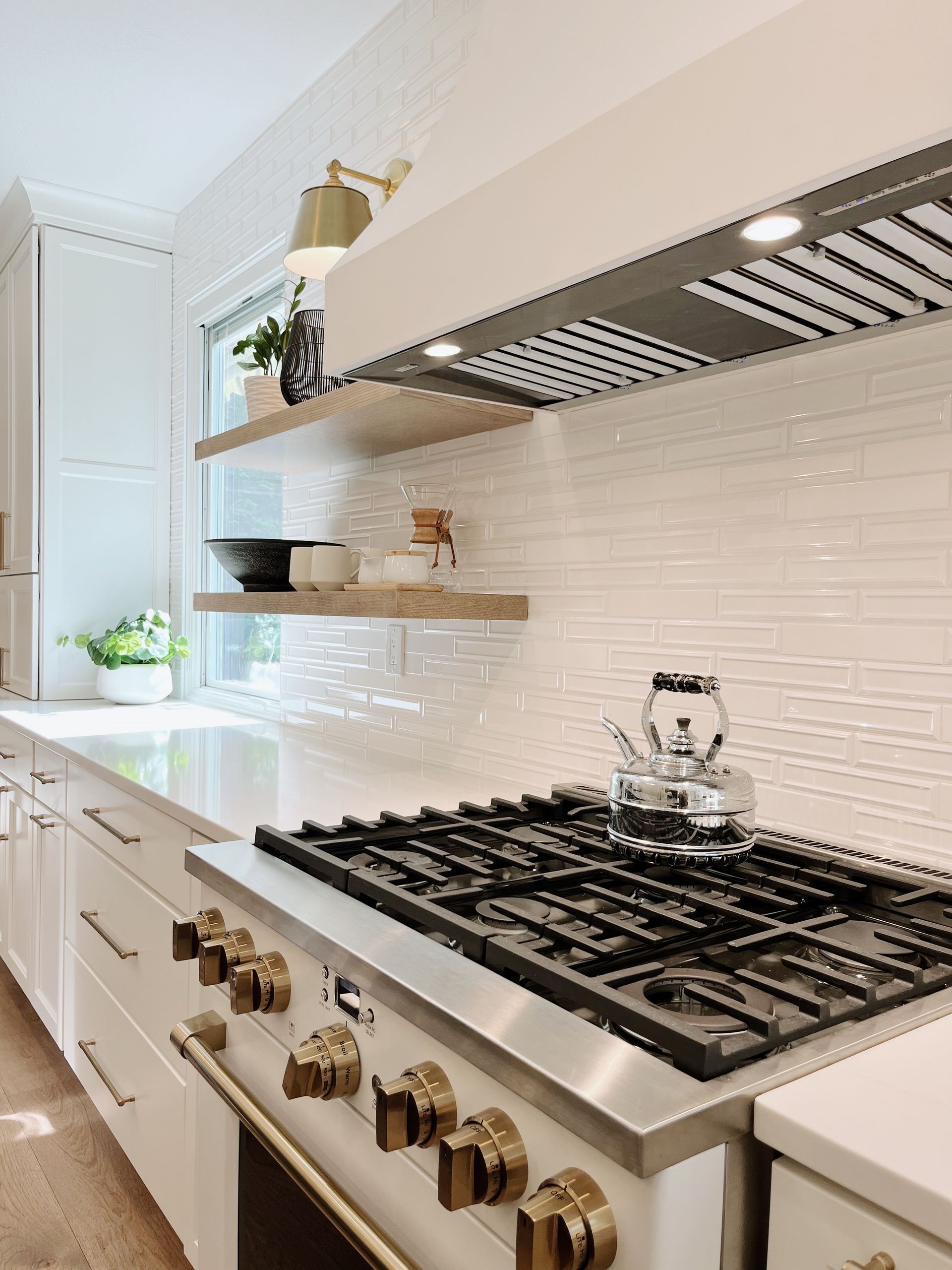 Bright white kitchen with stainless range, brass knobs, subway tile backsplash, and open shelving.