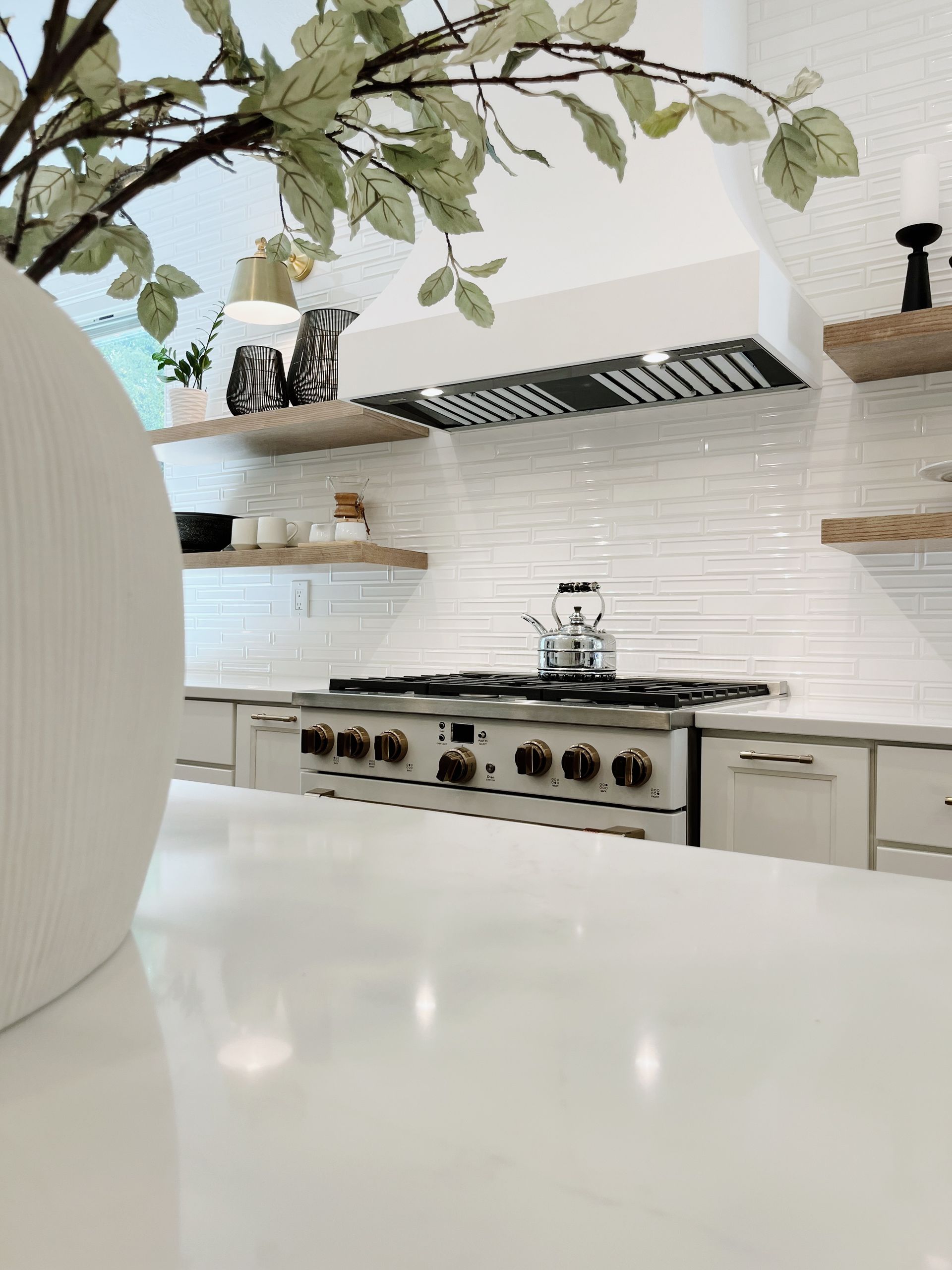 Bright modern kitchen with white tile backsplash, stainless steel stove, and wooden shelves under leafy branches