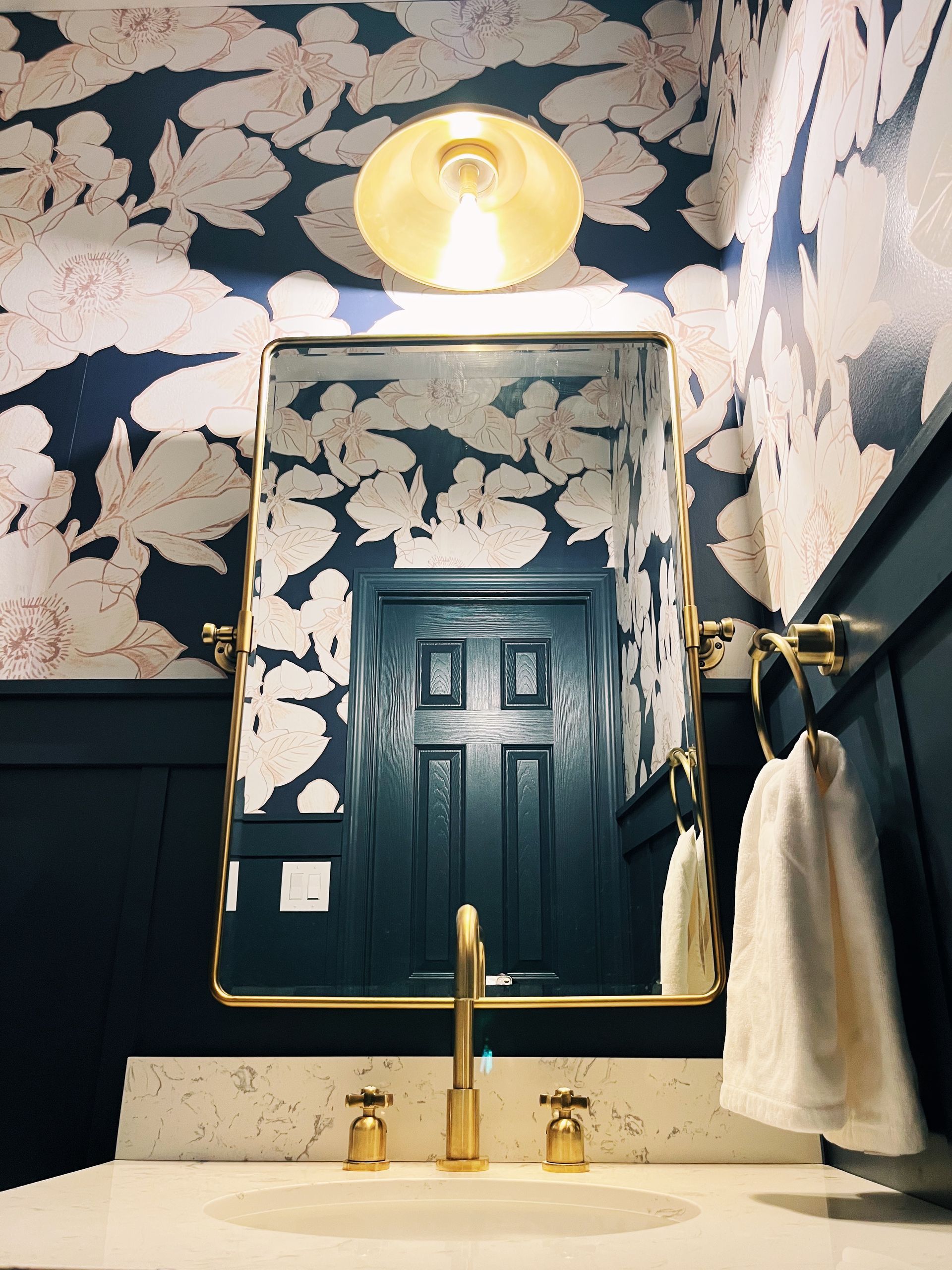 Dark navy floral bathroom with gold mirror, light fixture, faucet, and black door reflected in the mirror