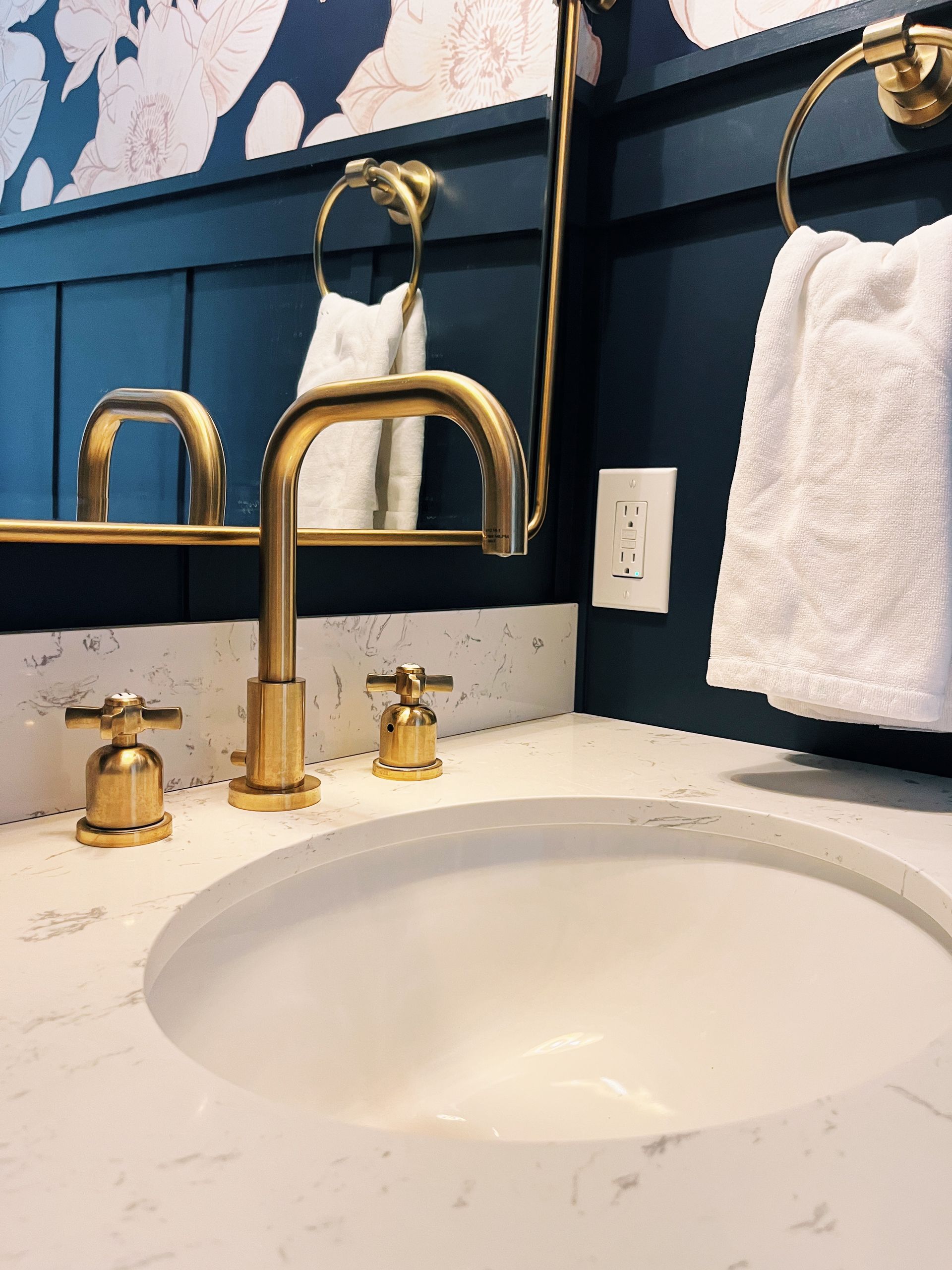 White bathroom vanity with oval sink, gold faucet, dark blue walls, and towels on hooks.