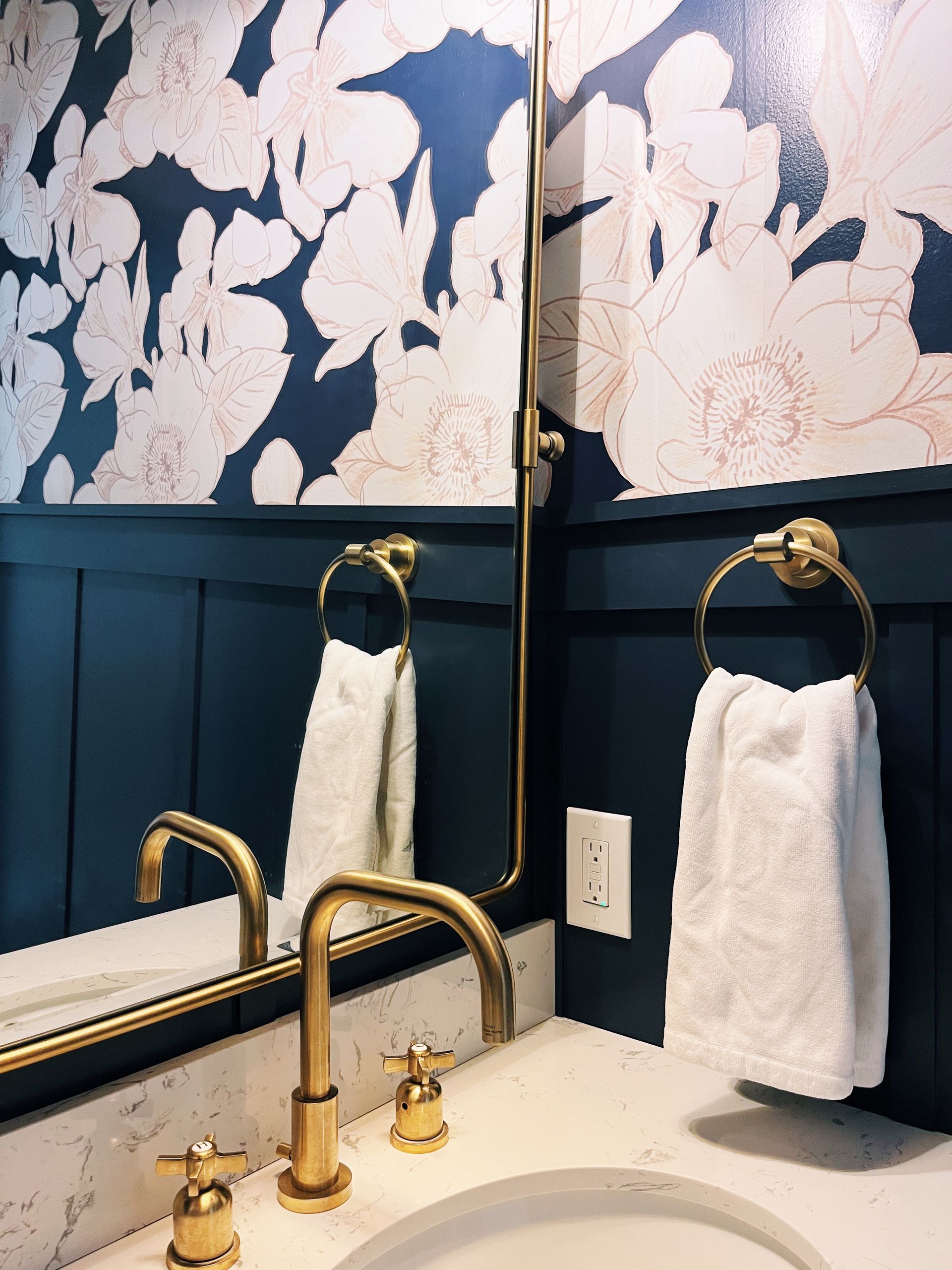 Bathroom sink with gold faucet, mirror, floral wallpaper, and white towels on navy walls