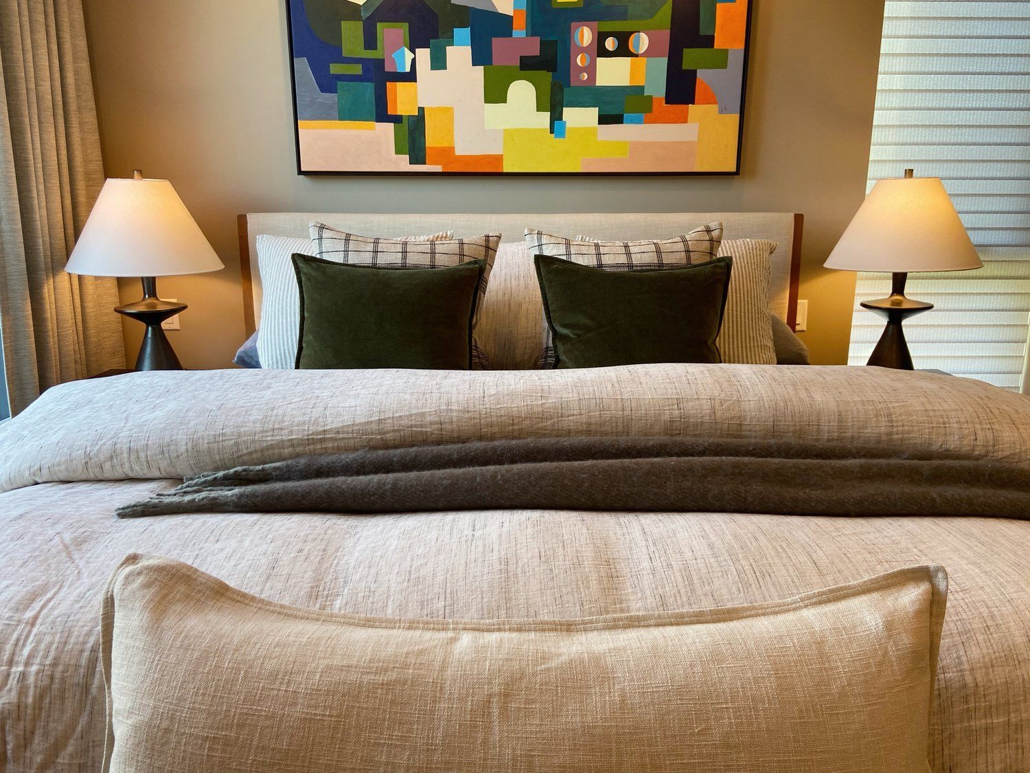A neatly made bed with gray bedding, bedside lamps, and colorful abstract wall art above the headboard.