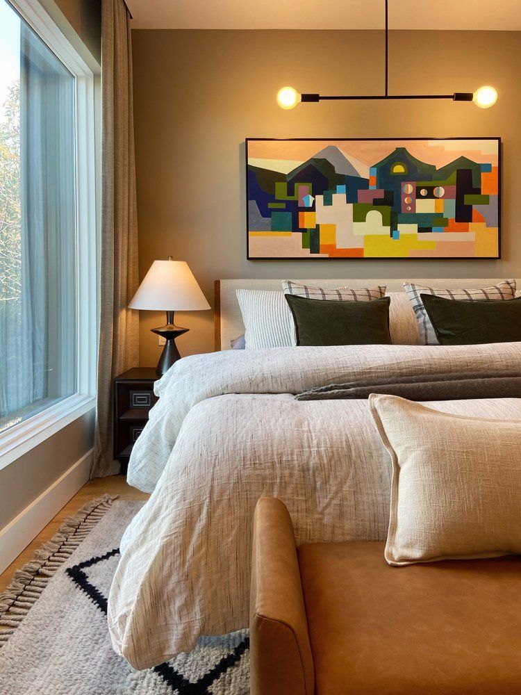 Cozy bedroom with a bed, tan chair, lamp, and colorful abstract artwork above the headboard.