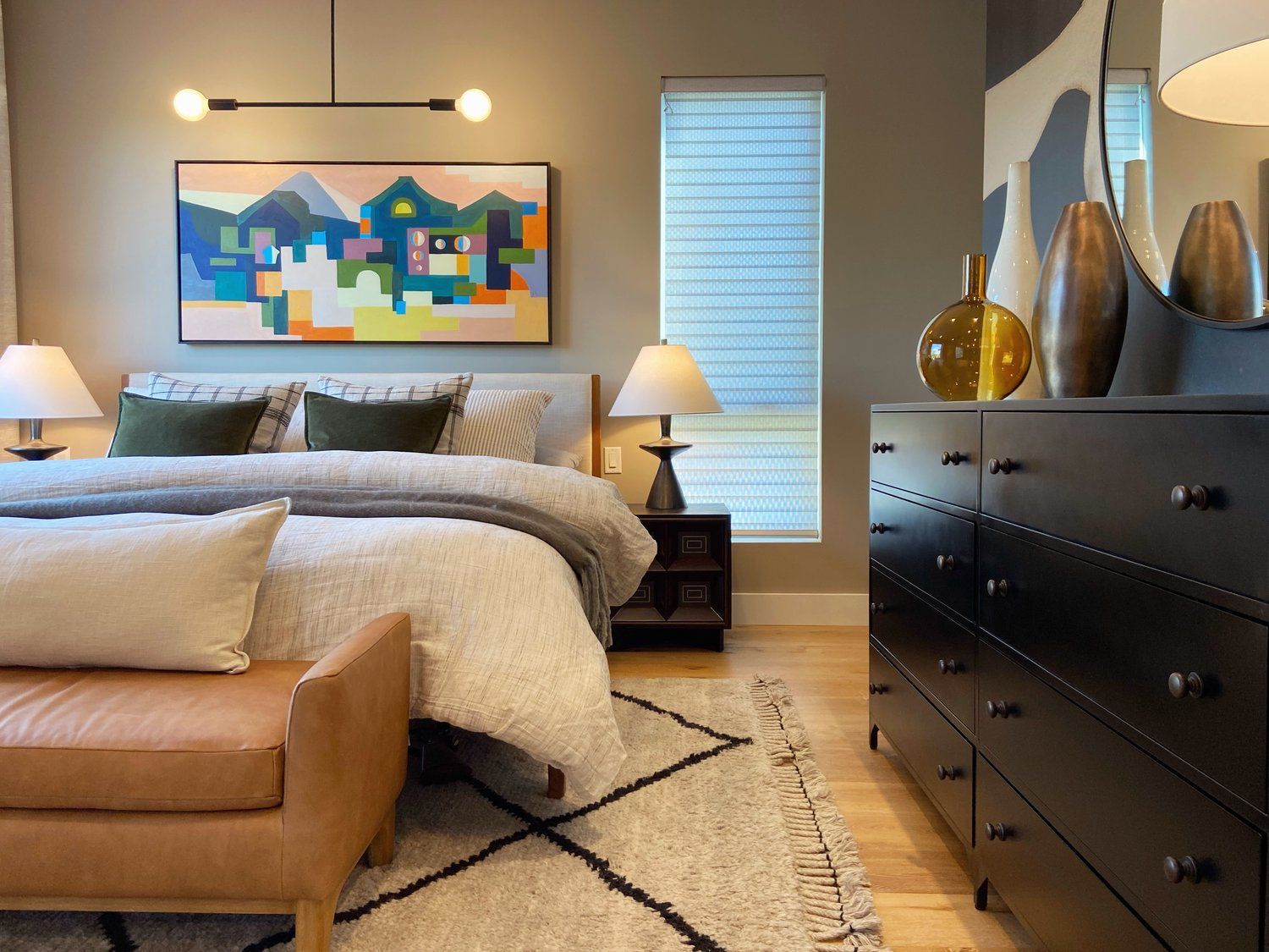 Cozy bedroom with a bed, tan bench, colorful wall art, lamps, and a dark dresser beside a window.
