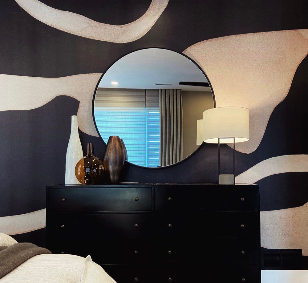 Black dresser with round mirror, lamp, and vases against abstract black-and-white wallpaper.