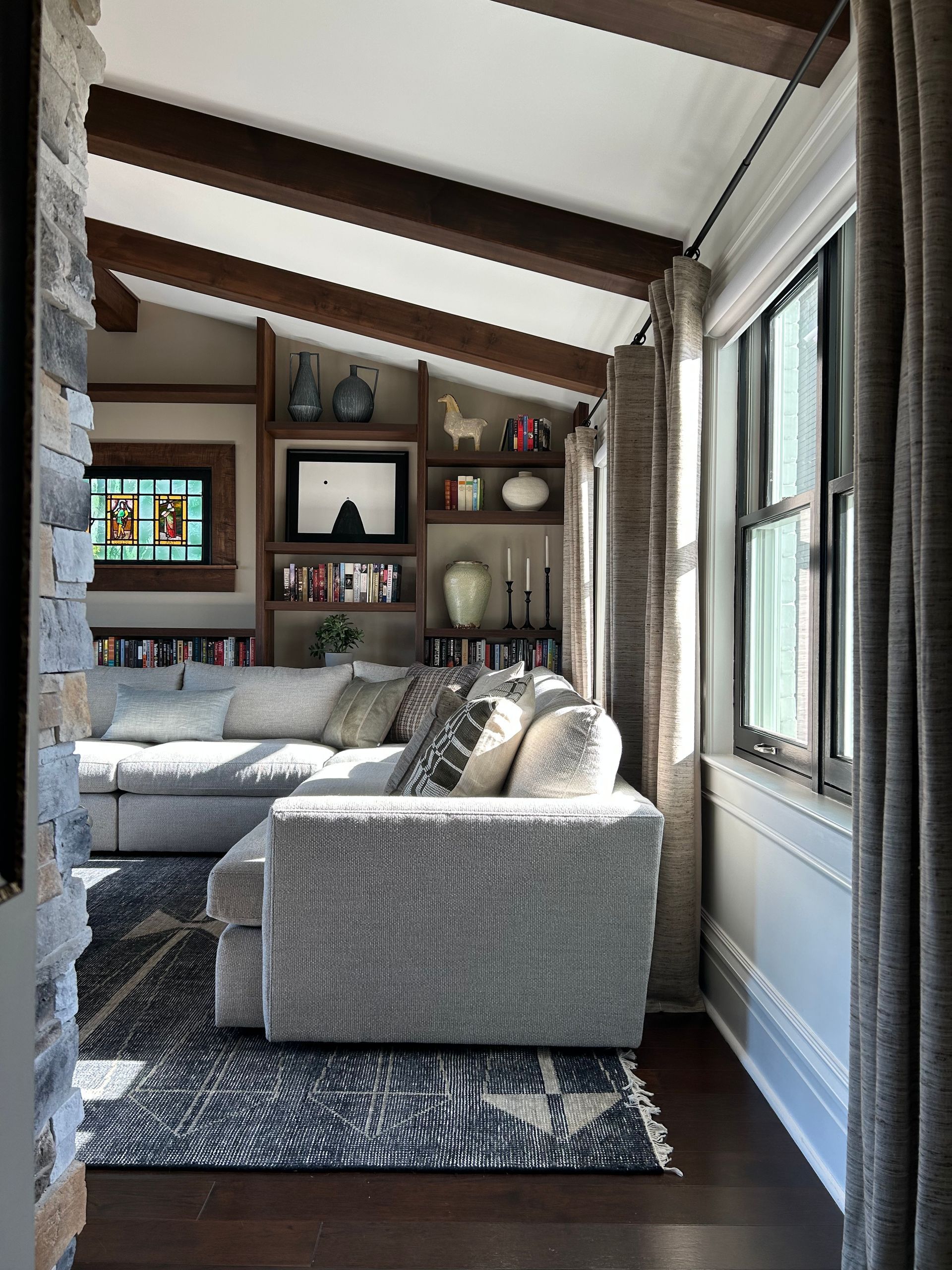 Cozy living room with gray sectional sofa, dark rug, wood beams, and bright windows